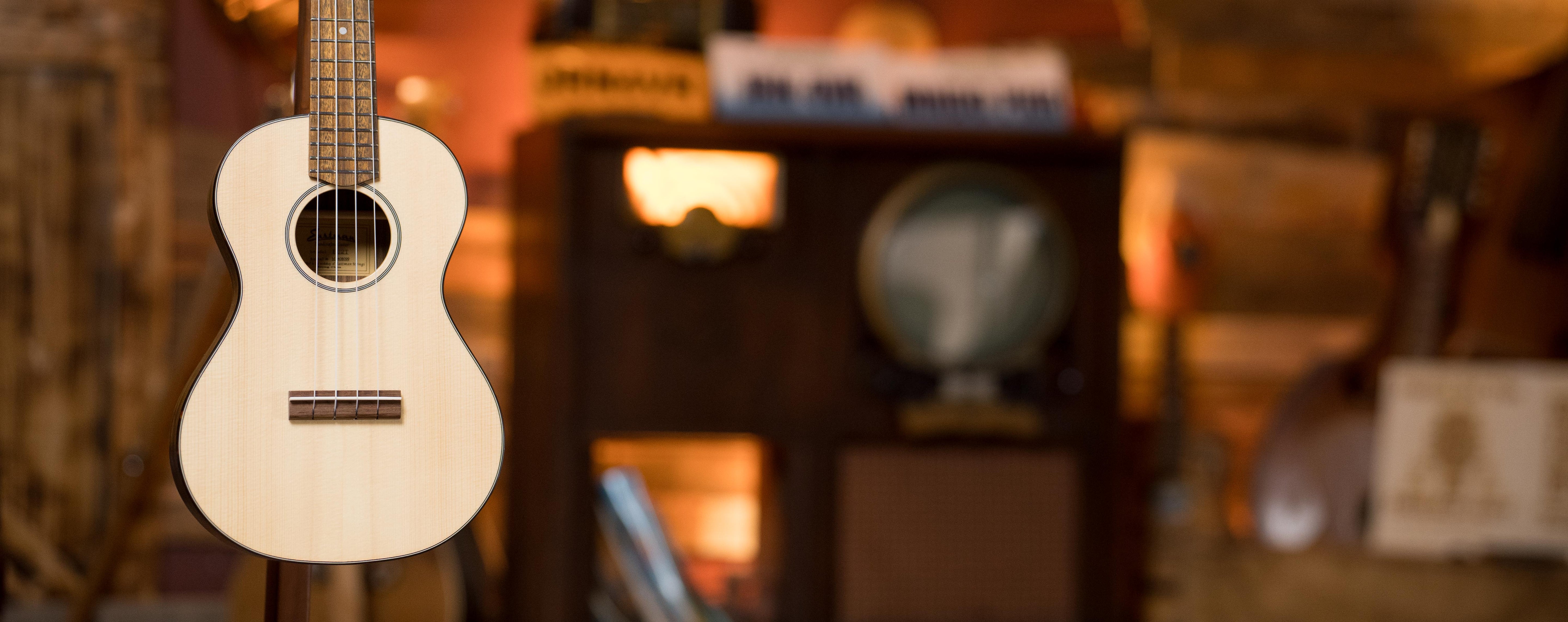A close-up of a light-colored ukulele hanging in front of a blurred background with vintage furniture and warm lighting.