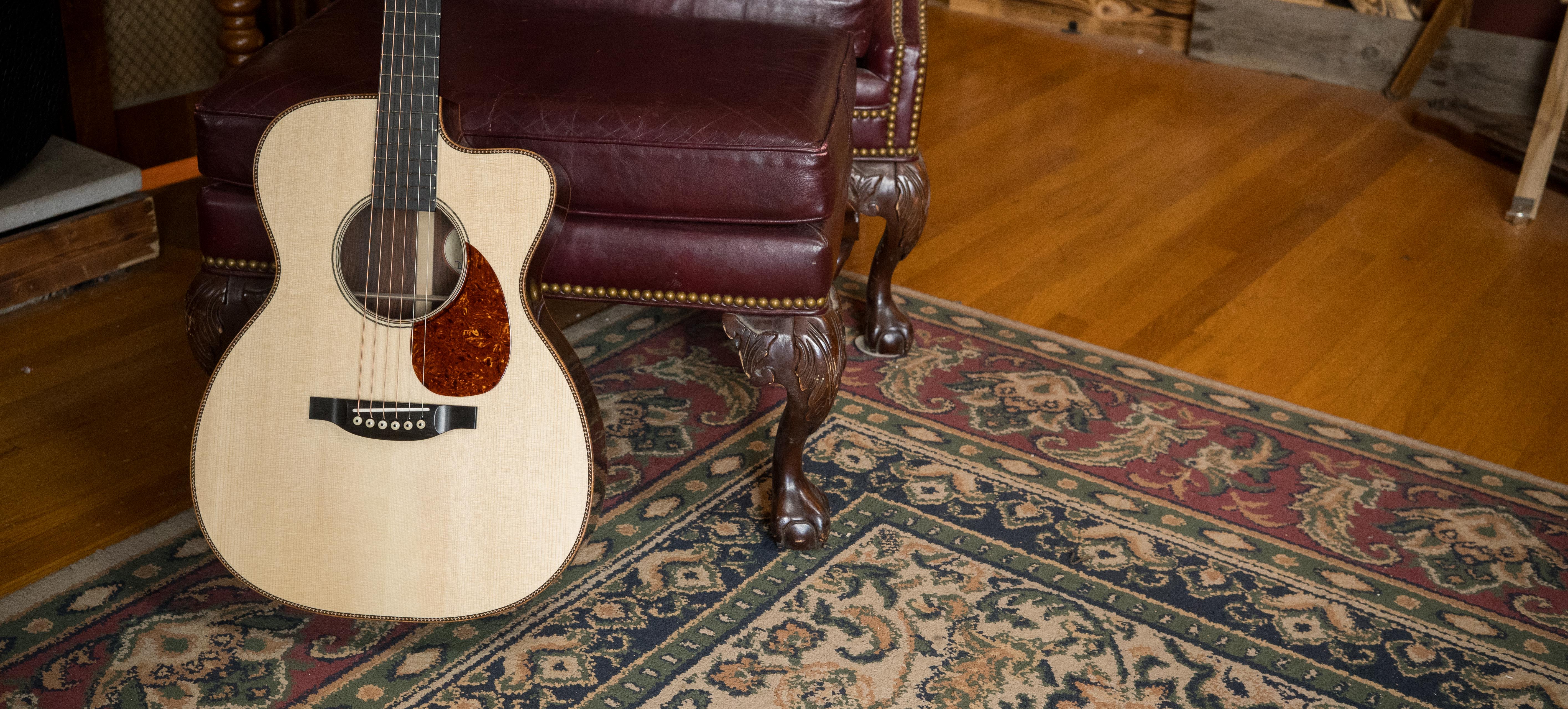 A light-colored acoustic guitar leans against a burgundy leather chair with ornate wooden legs, placed on a patterned rug over a hardwood floor in a cozy room.