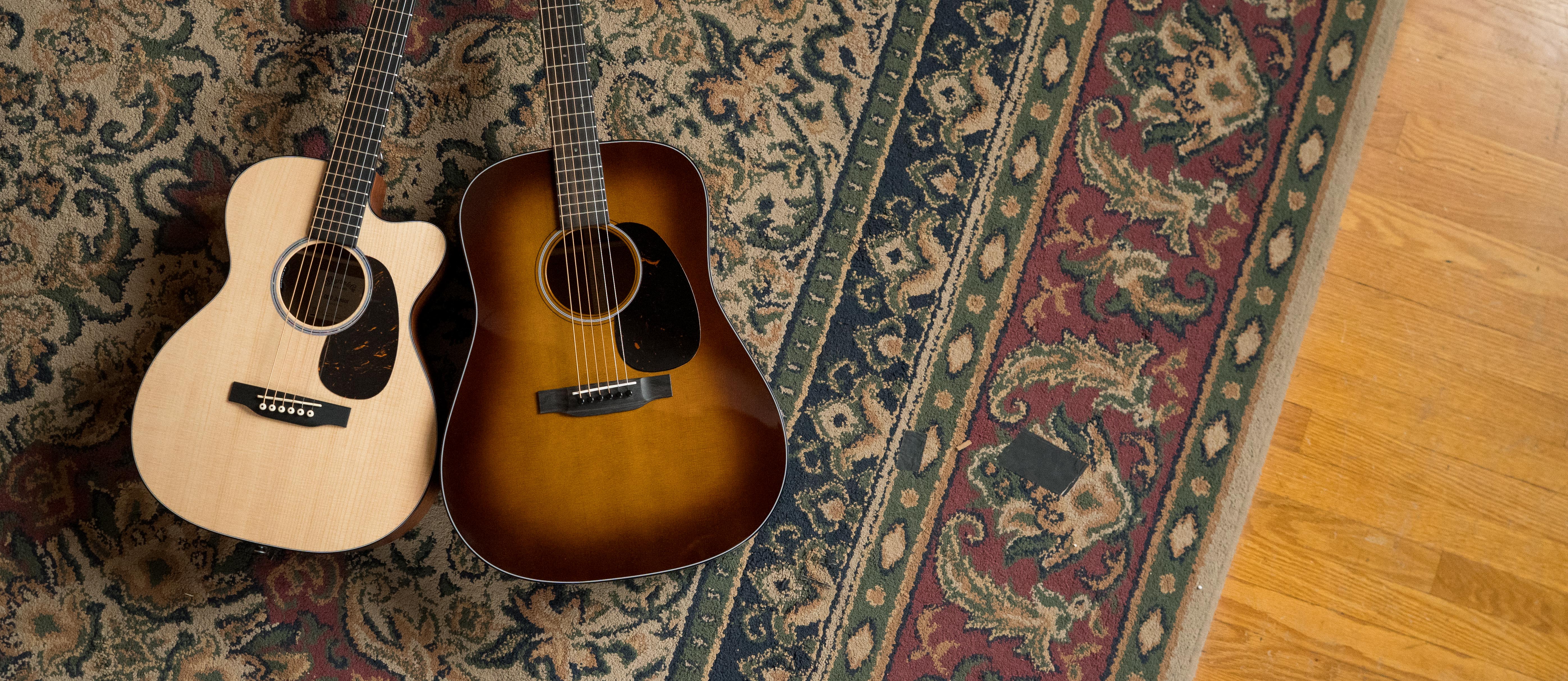 Two acoustic guitars, one with a light wood finish and one with a sunburst finish, rest side by side on a patterned rug next to a hardwood floor.