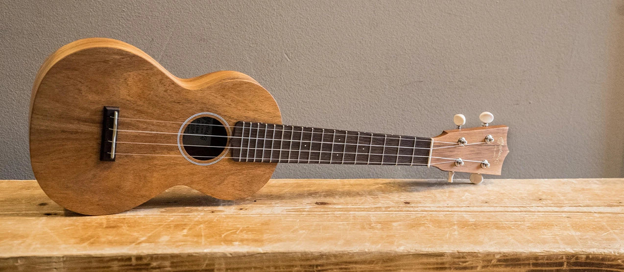 A wooden ukulele with a dark fretboard rests on a rustic wooden surface against a plain gray wall.