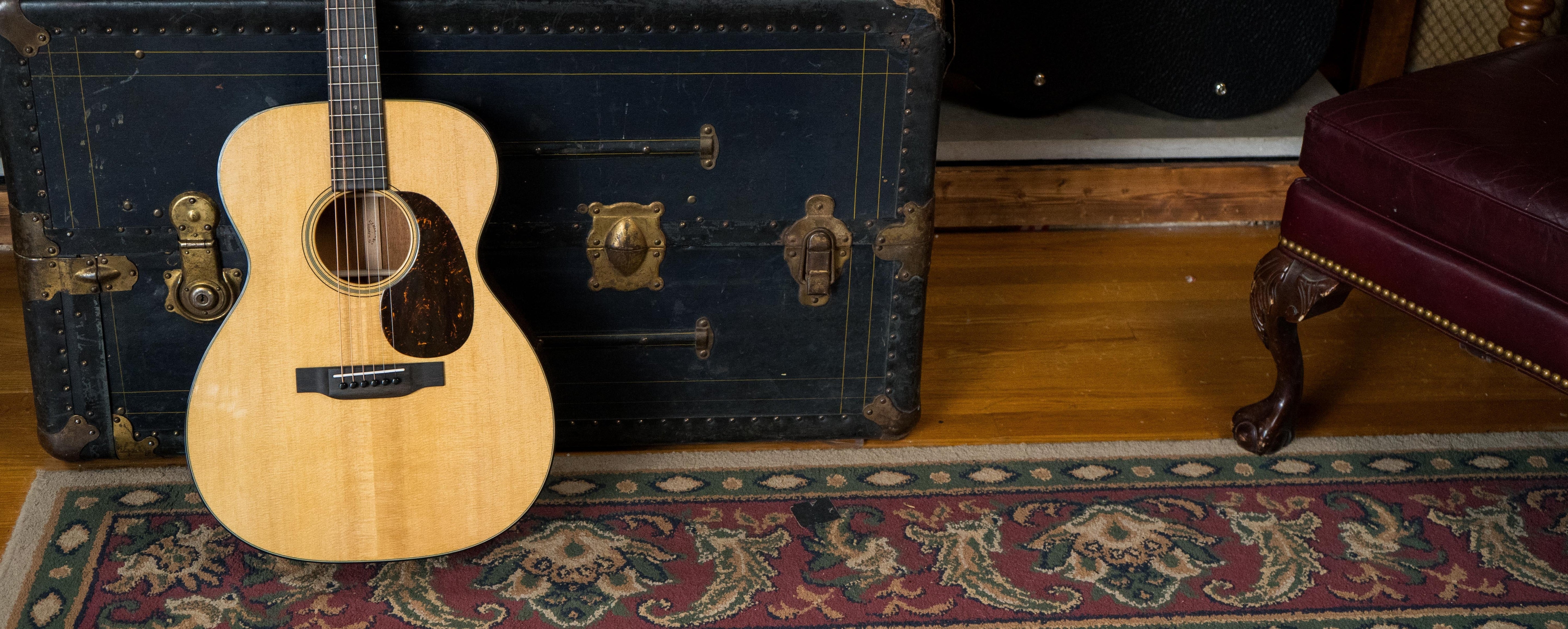 An acoustic guitar leans against a vintage black trunk on a wooden floor, next to a decorative rug and a burgundy leather chair with carved wooden legs.