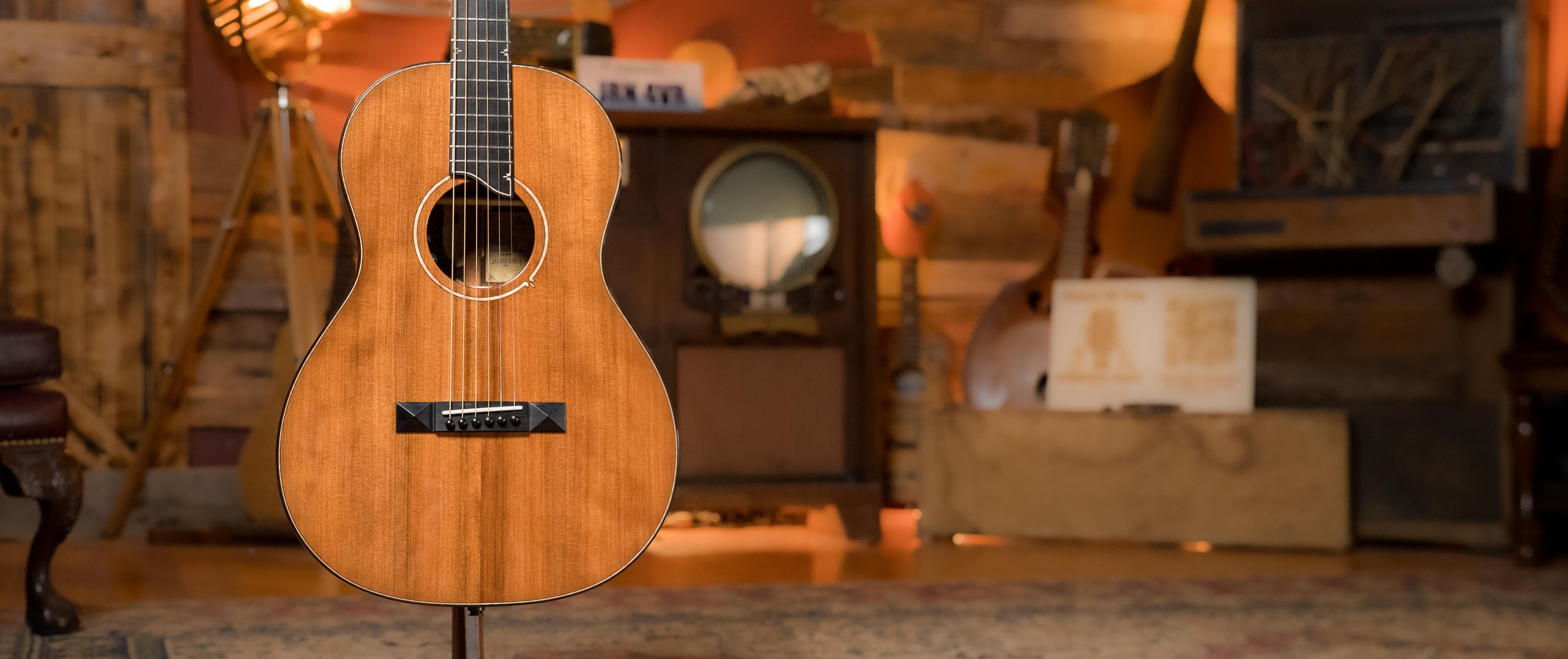 An acoustic guitar with a wooden finish is displayed upright in a cozy, rustic room with vintage decor and warm lighting.