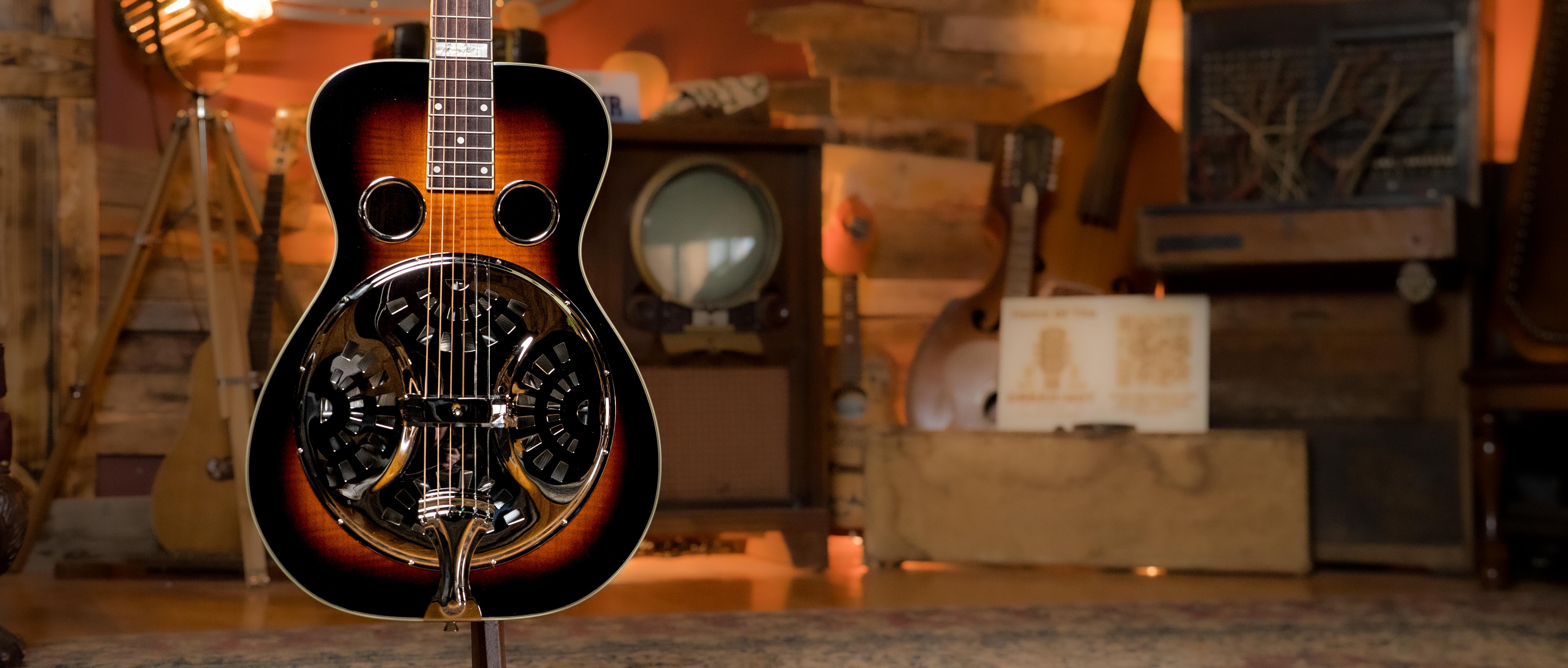 A resonator guitar with a sunburst finish is displayed on a stand in a cozy room with vintage decor, including an old television, wooden panels, and various musical instruments in the background.
