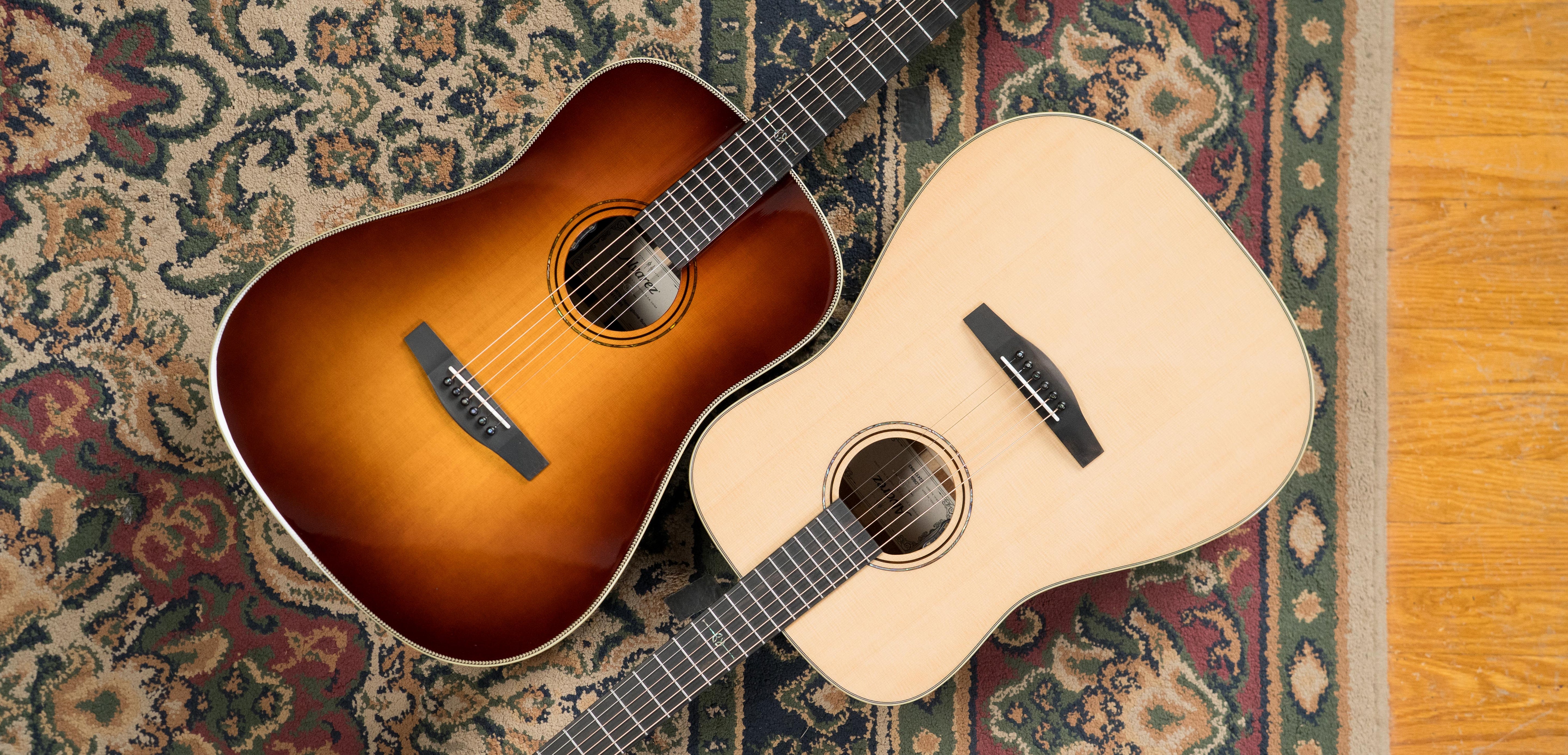 Two acoustic guitars, one with a sunburst finish and one with a natural wood finish, lying side by side on a patterned rug with a border next to a hardwood floor.