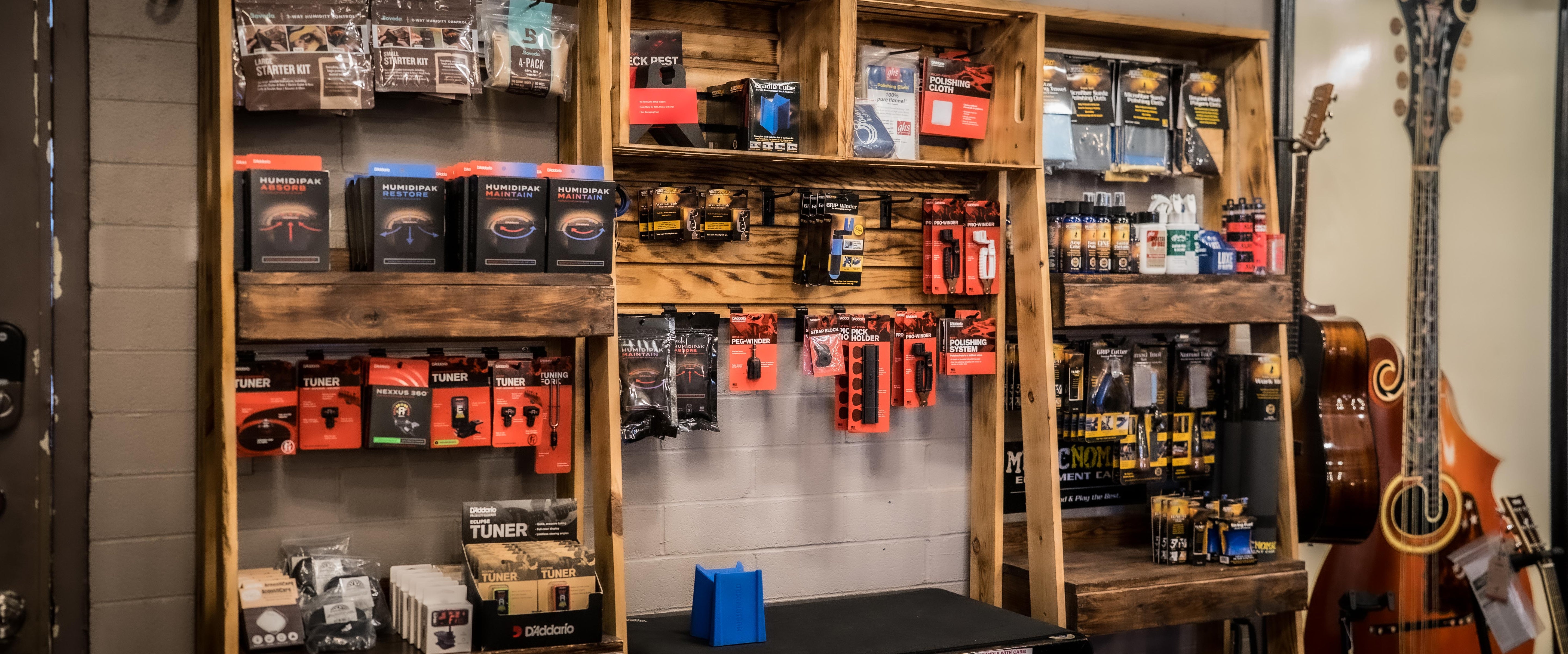 Wall-mounted shelves display various music accessories like guitar strings, tuners, picks, and capos in colorful packaging. An acoustic guitar and double bass are visible on the right side.