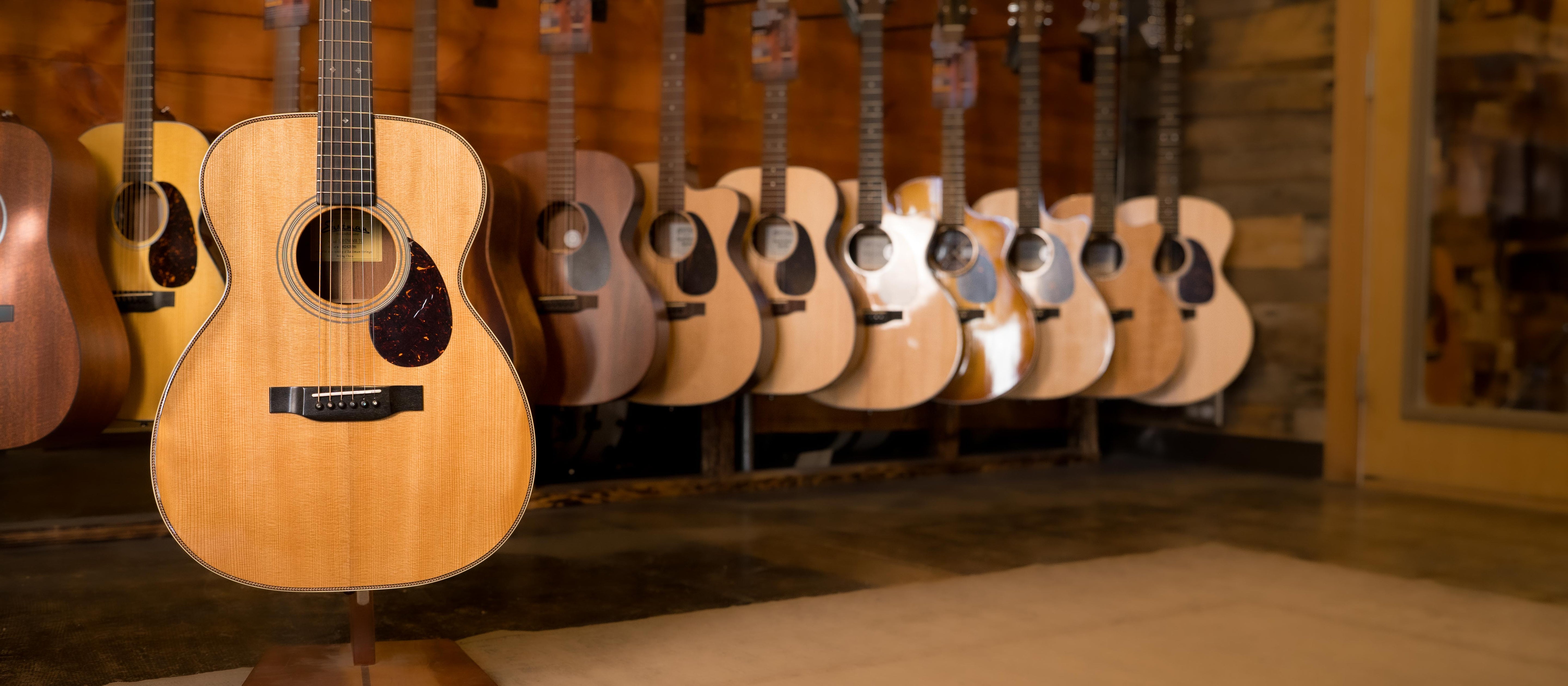 An acoustic guitar is displayed on a stand in the foreground, with several other acoustic guitars hanging on a wooden wall in the background inside a music store.