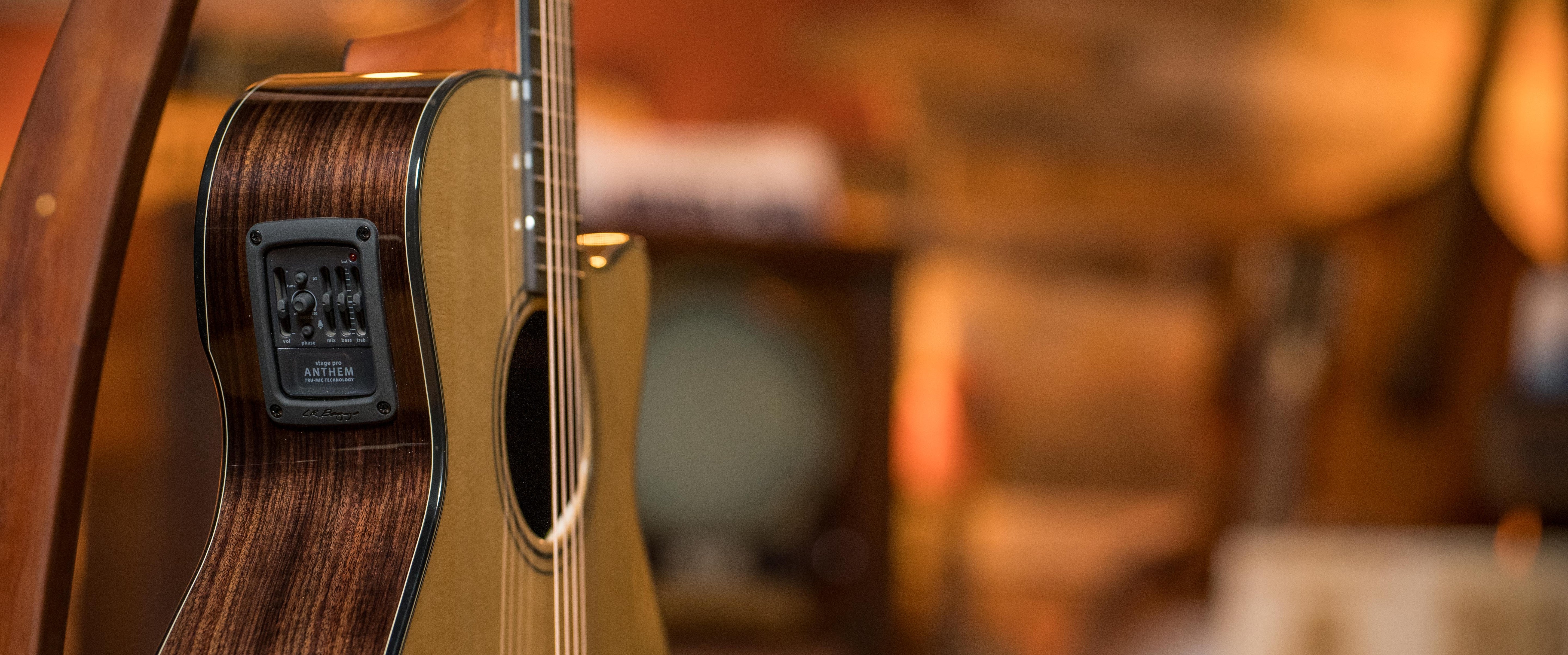 Close-up of an acoustic guitar with control panel on its side, focusing on the wooden grain and strings; blurred background with warm lighting and indistinct objects.