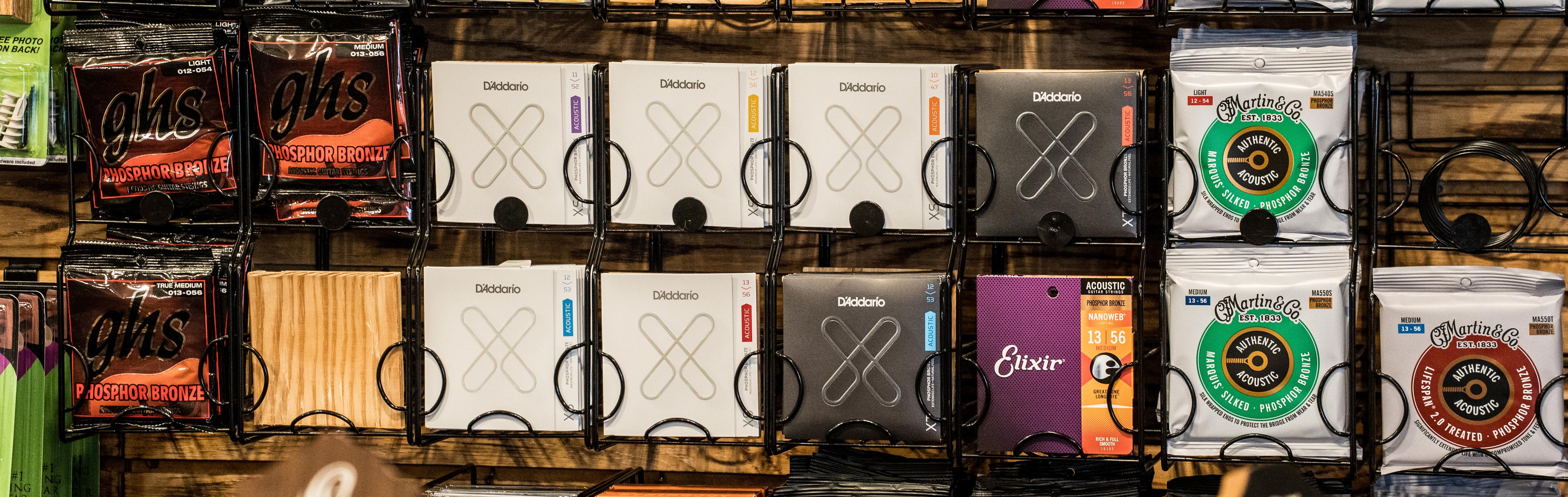 A display rack holds various packs of guitar strings, including brands like D’Addario, Elixir, and Ernie Ball, organized neatly in rows at a music store.