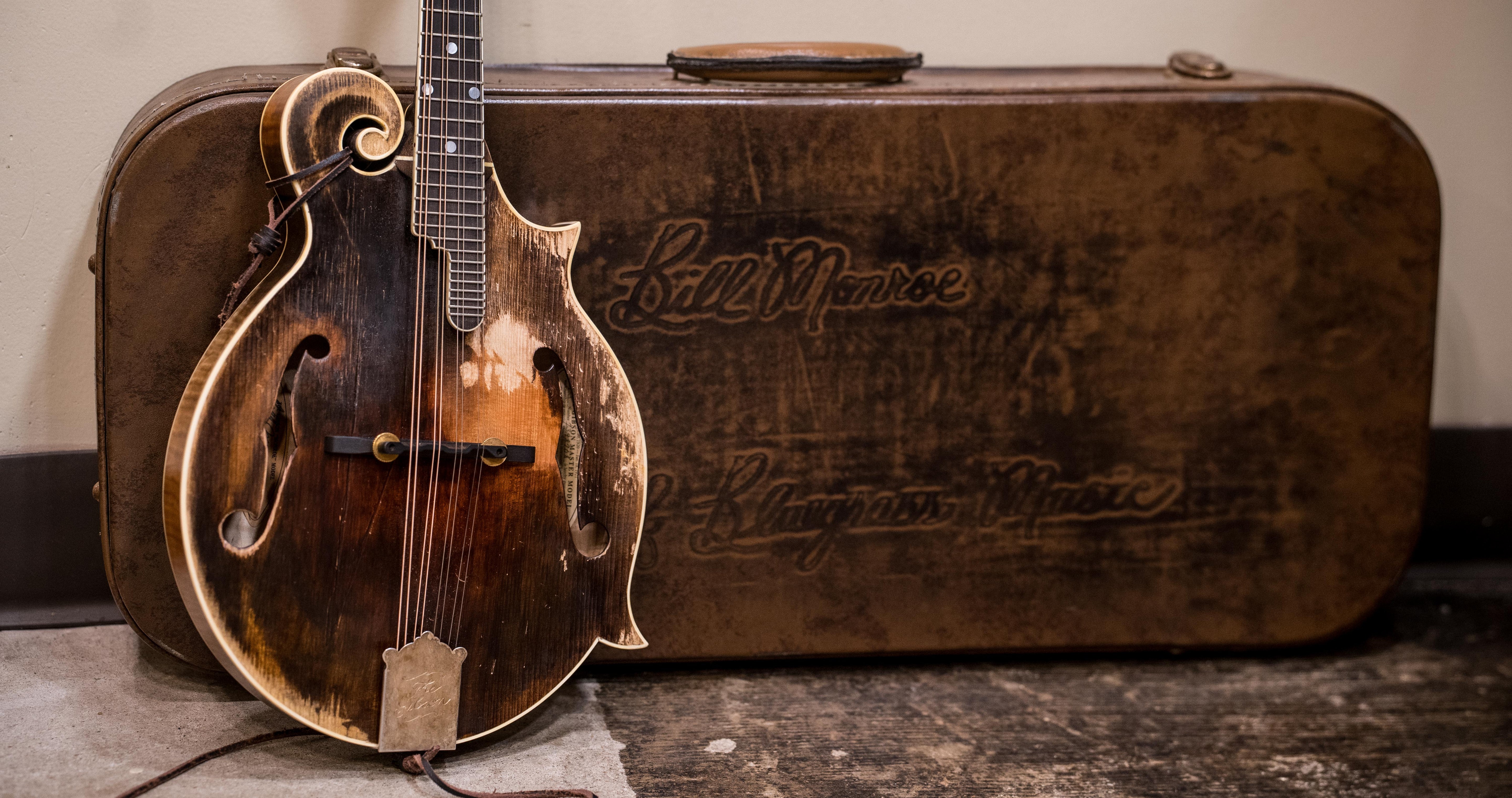 A worn mandolin rests against a vintage, weathered brown case embossed with “Bill Monroe” and “Bluegrass Music.”.