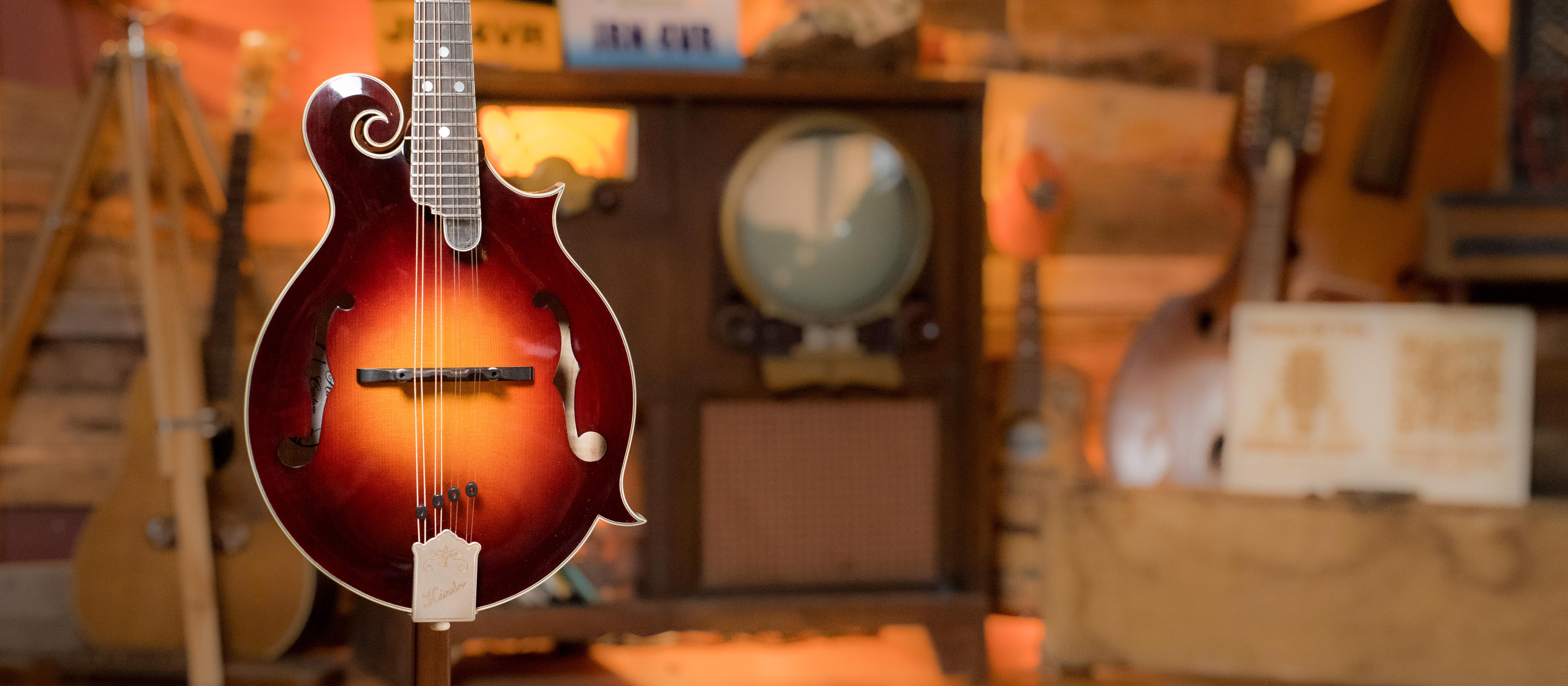 A mandolin with a sunburst finish hangs in the foreground, with a vintage television and other musical instruments in a warmly lit, rustic room in the background.