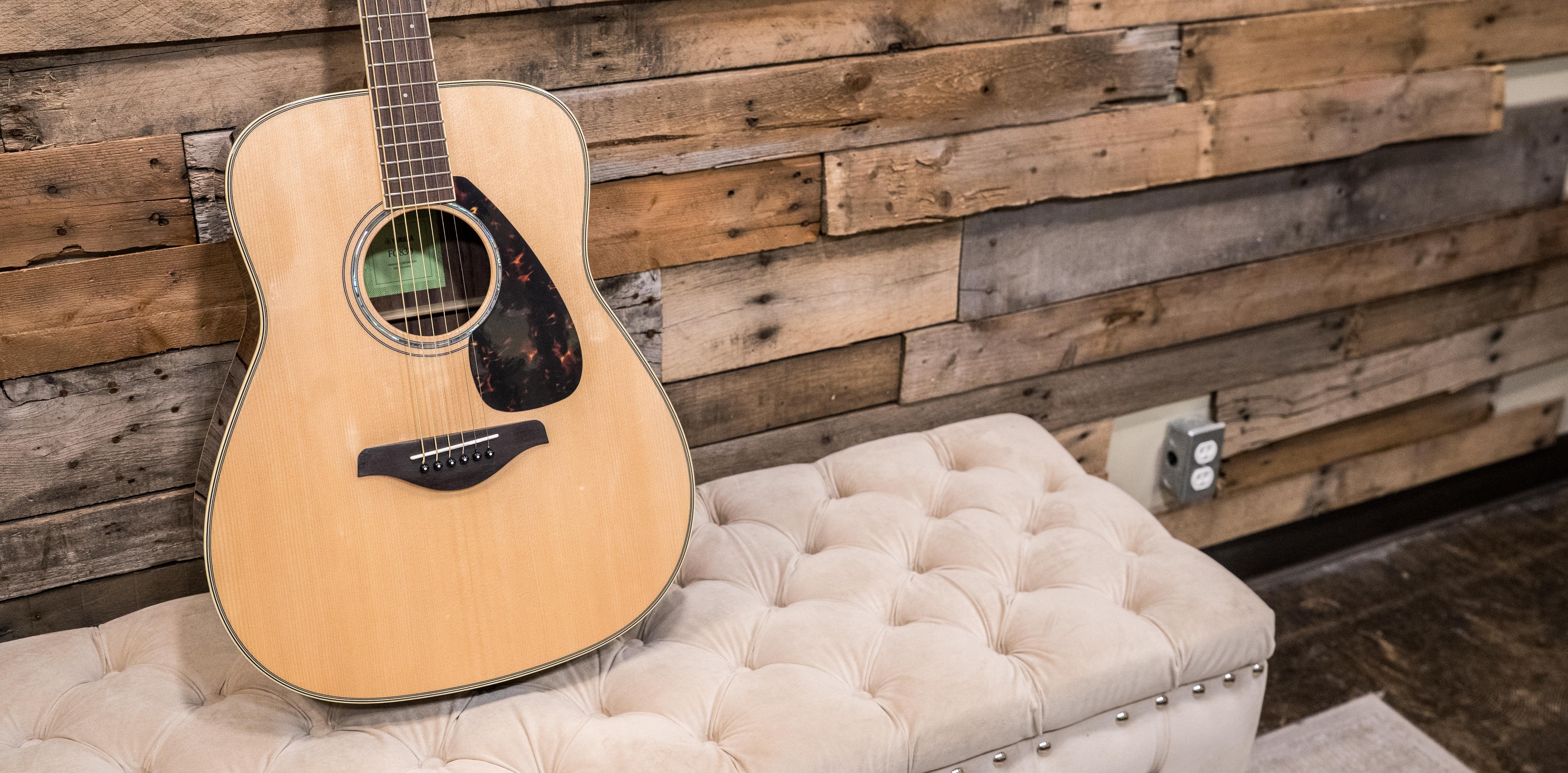 An acoustic guitar rests on a tufted cream-colored bench in front of a rustic wooden plank wall with an electrical outlet visible on the right.