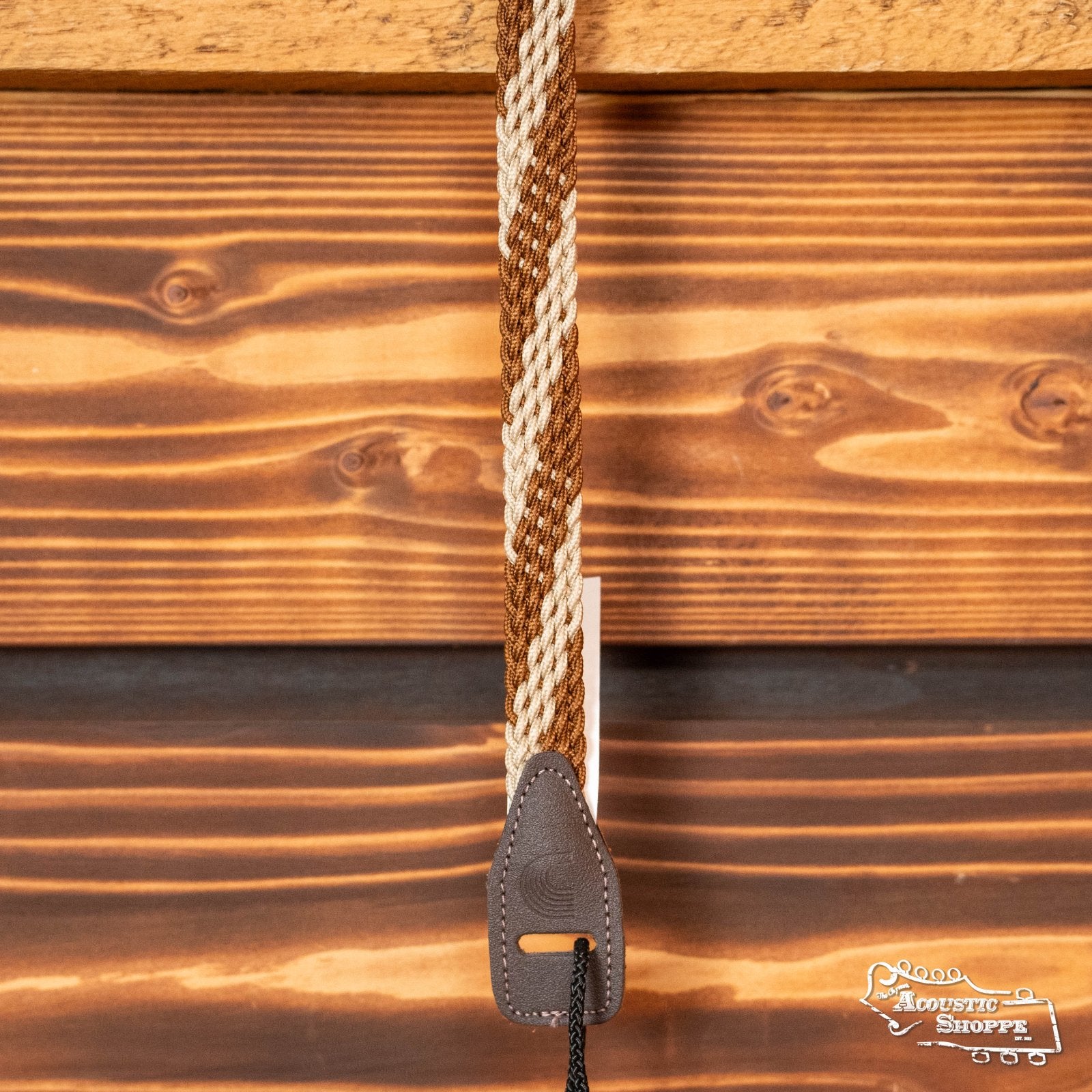 The DAddario 10MB00 Braided Leather 1 Mandolin Strap in brown/cream hangs against a wooden wall. Its stitched leather end with attachment string is shown, with The Acoustic Shoppe logo in the bottom right corner.