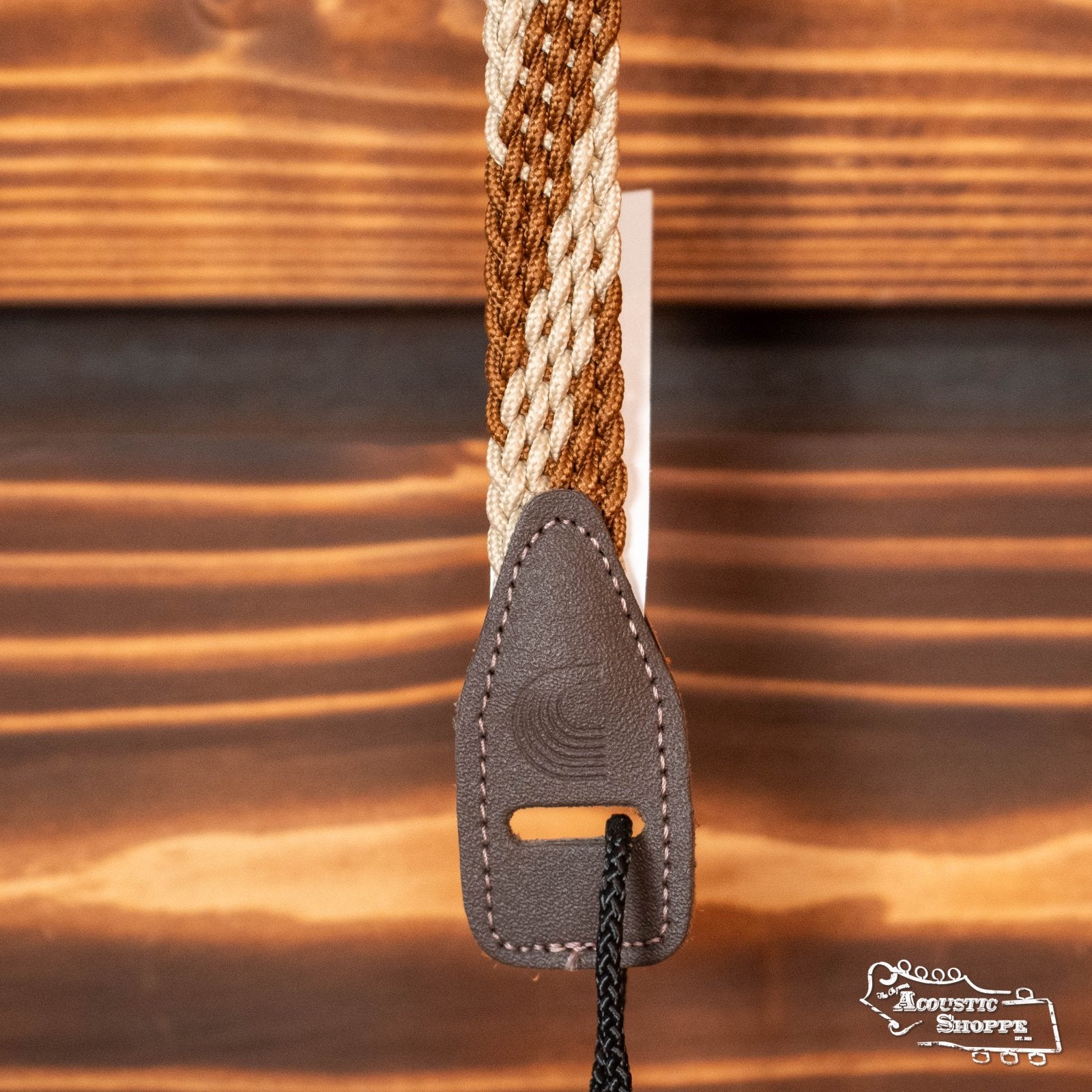 A close-up of the DAddario 10MB00 Braided Leather 1 Mandolin Strap in brown/cream, featuring braided brown and beige fabric with a dark brown leather end, set on a wooden background. The Acoustic Shoppe logo appears at the bottom right.
