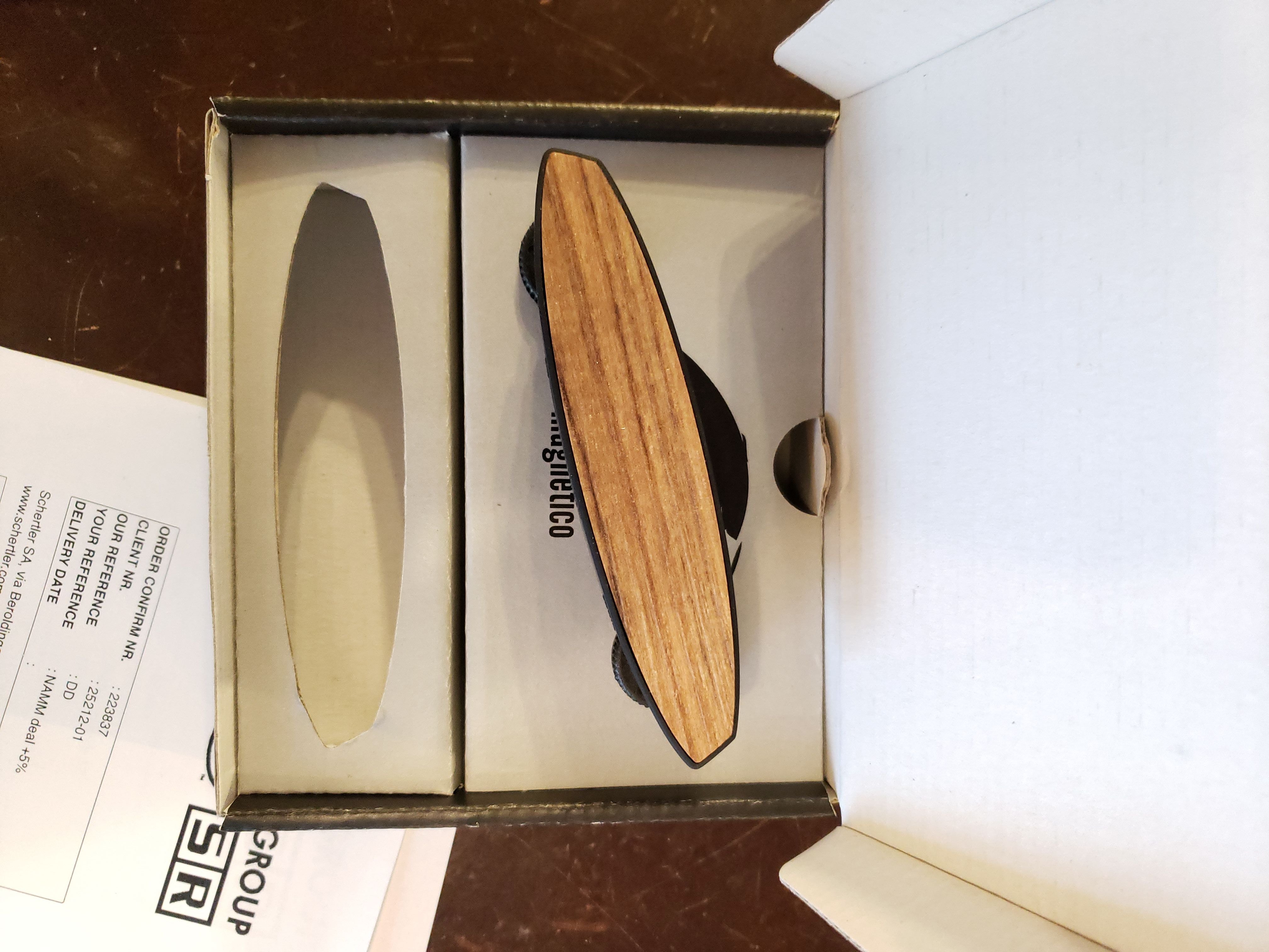 A Schertler Magnetico M-AG6-LIMITED acoustic guitar pickup (light wood top) by The Acoustic Shoppe rests in an open box, with a printed paper beside it, reminiscent of the careful packaging for fine musical gear.