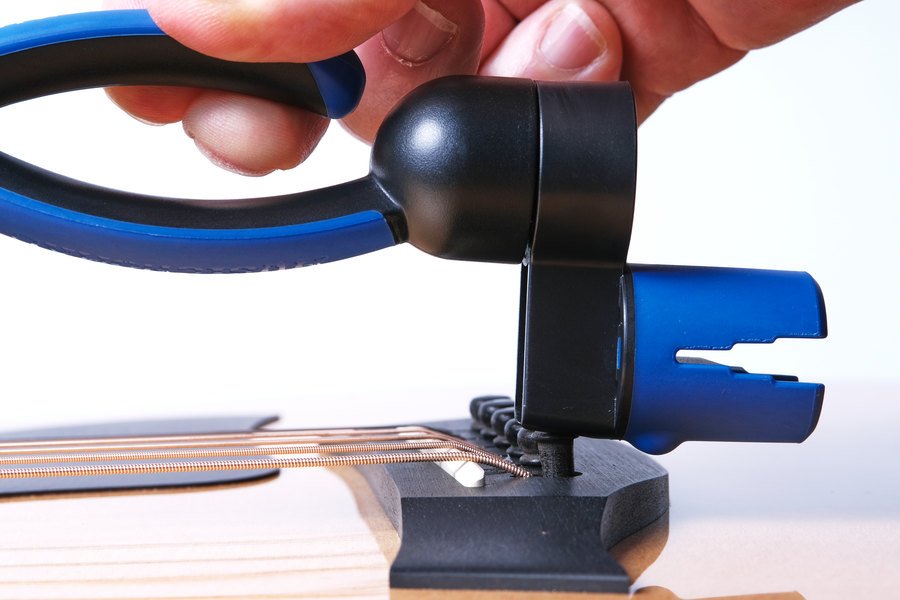 A hand uses the Music Nomad GRIP ONE (MN223), an all-in-one string winder, cutter, and puller, to cut or bend strings on the bridge of an acoustic guitar.