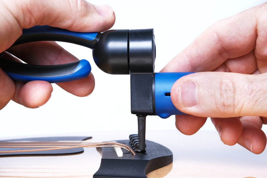 Close-up of hands using the Music Nomad GRIP ONE - All in ONE String Winder, Cutter, Puller (MN223) to wind guitar strings onto tuning pegs; headstock and several metal strings are visible. Brand: Music Nomad.