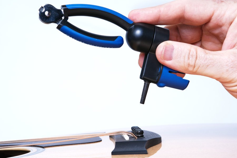 A hand holds the Music Nomad GRIP ONE (MN223) All in ONE String Winder, Cutter, and Puller above an acoustic guitar bridge, ready to restring or remove a string using this Music Nomad tool.