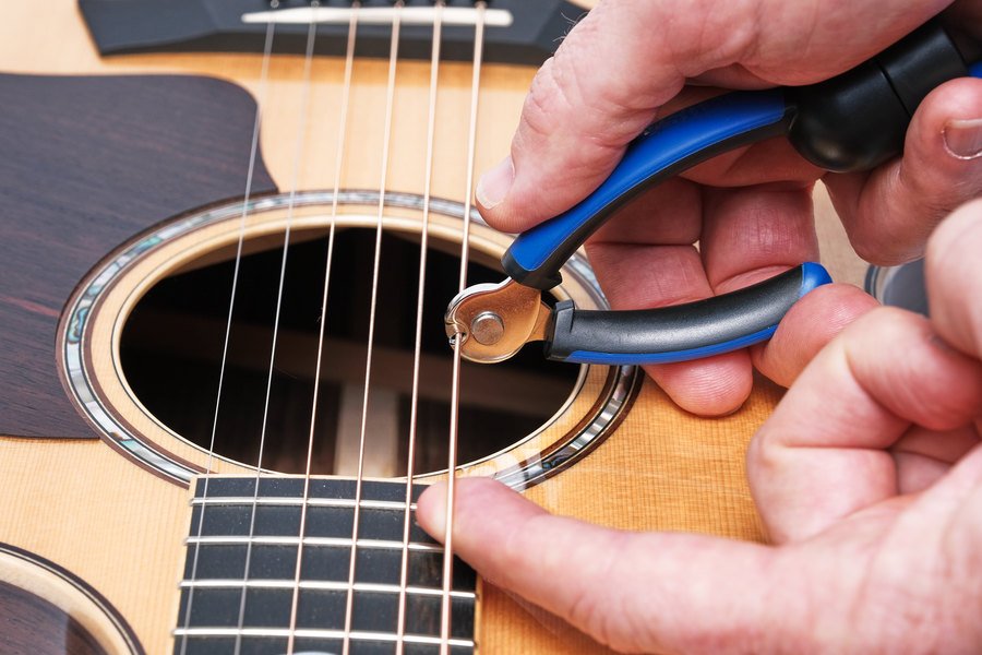Close-up of hands using the Music Nomad GRIP ONE (MN223), an all-in-one string winder, cutter, and puller, to trim guitar strings near the sound hole of an acoustic guitar.