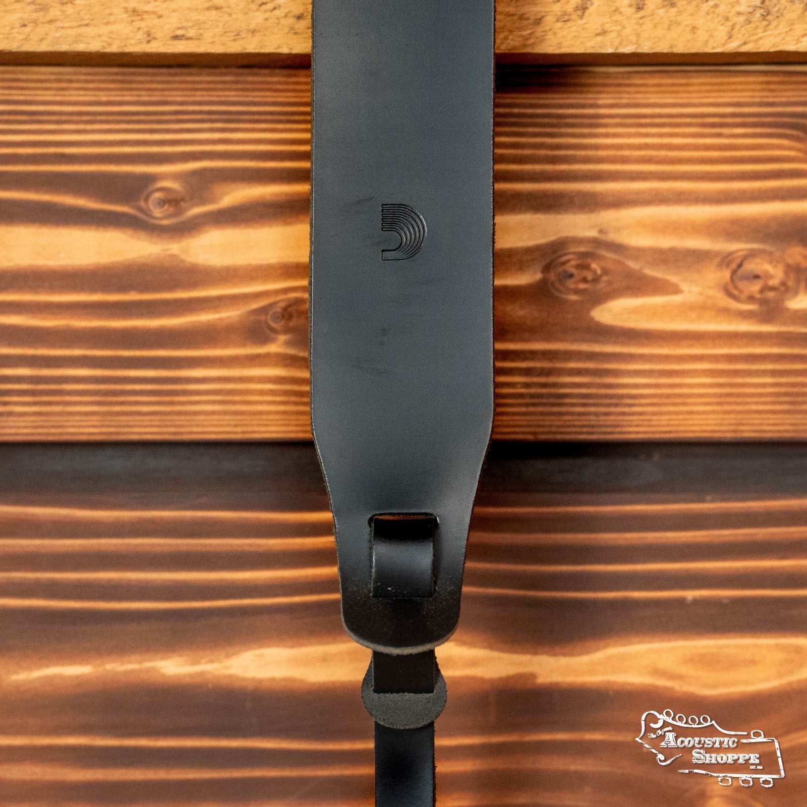 The DAddario 25LBNJ00-DX Classic 2 Leather Banjo Strap in black hangs against a wooden backdrop, featuring a subtle embossed logo. Adjustable for comfort and style, itโs ideal for any musician. The Acoustic Shoppe logo appears bottom right.