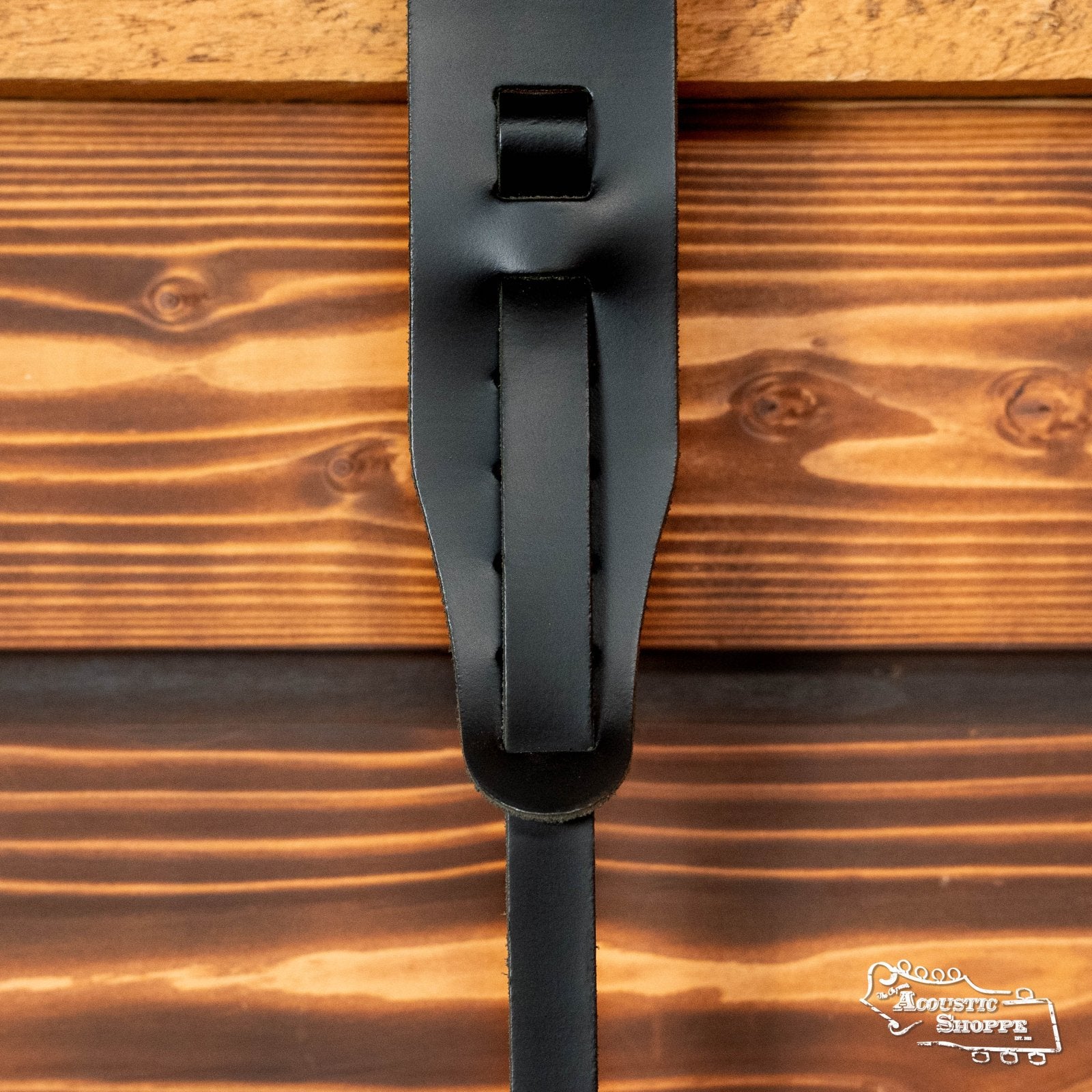 Close-up of a DAddario 25LBNJ00-DX Classic 2 Leather Banjo Strap - Black, mounted on varnished wooden planks. The Acoustic Shoppe logo appears in the lower right corner, adding a rustic touch.