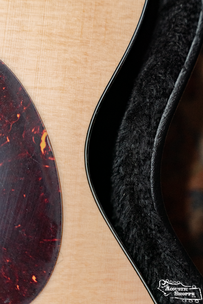 A close-up of an acoustic guitar with a light wood finish and dark tortoiseshell pickguard beside the Guardian CG-022-OOO Deluxe Hardshell Case-000 Style by Guardian, featuring a plush black lining. Available now at Acoustic Shoppe.