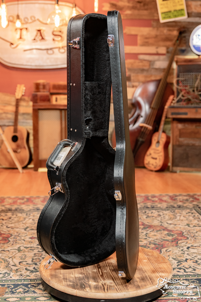 A Guardian CG-022-OOO Deluxe Hardshell Case-000 Style by Guardian stands open and upright on a wooden platform in a cozy, vintage-style music room at the Acoustic Shoppe, surrounded by guitars, a bass, and amplifiers.
