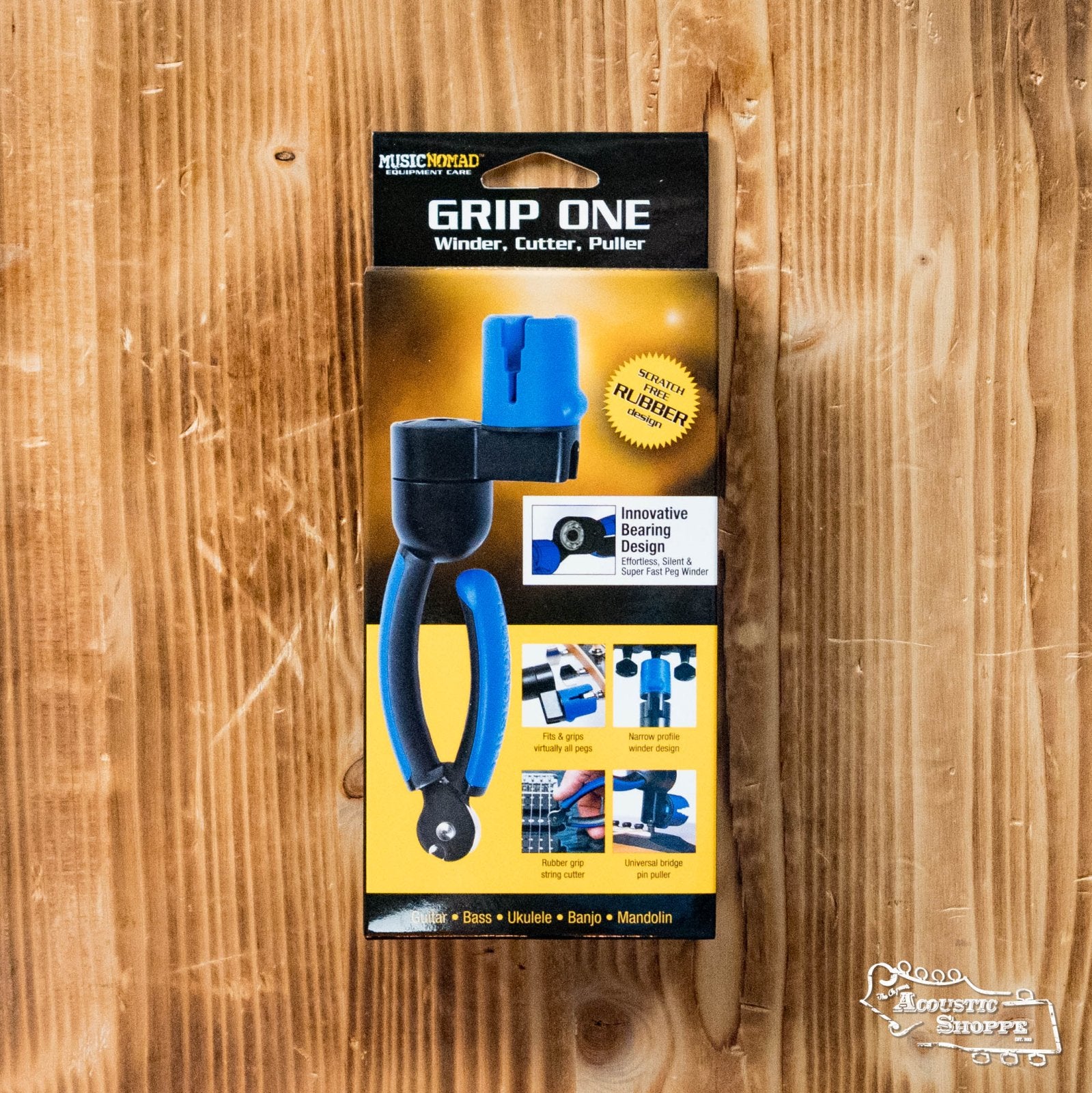 The Music Nomad GRIP ONE (MN223) All-in-ONE String Winder, Cutter, and Puller is shown in its blue and black packaging on a wooden surface. It works for guitar, bass, ukulele, banjo, and mandolin.