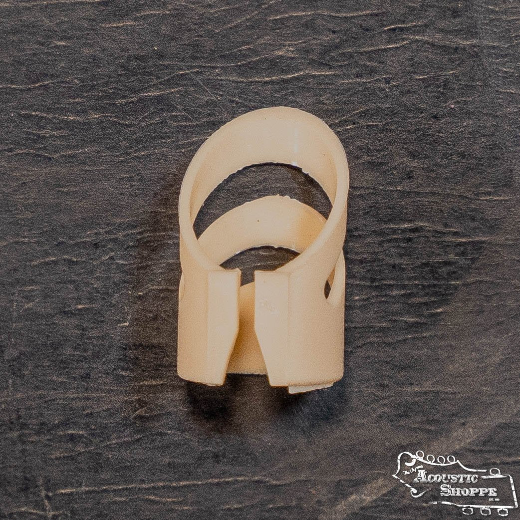 A large, beige plastic ring-shaped Alaska Picks ALP-12 finger pick by Alaska Pik, featuring a split design with overlapping ends, is placed on a dark textured surface. The Acoustic Shoppe logo appears in the bottom right corner.
