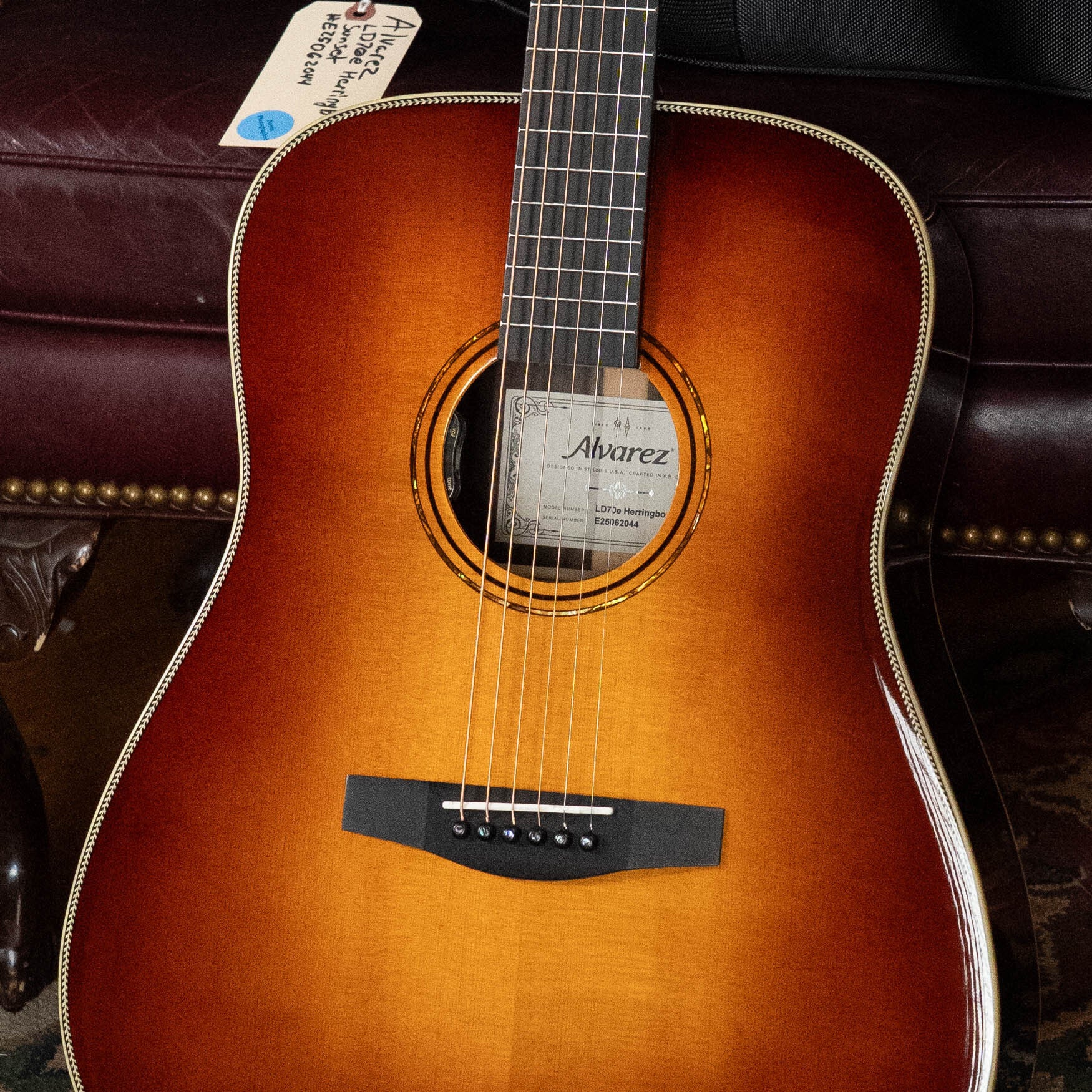 A close-up of the Alvarez Laureate Series LD70e Herringbone Sunset Sitka/Rosewood Dreadnought Guitar with LR Baggs Hifi Pickup shows its sunburst finish, strings, soundhole, and a tag near the headstock against a leather couch background.