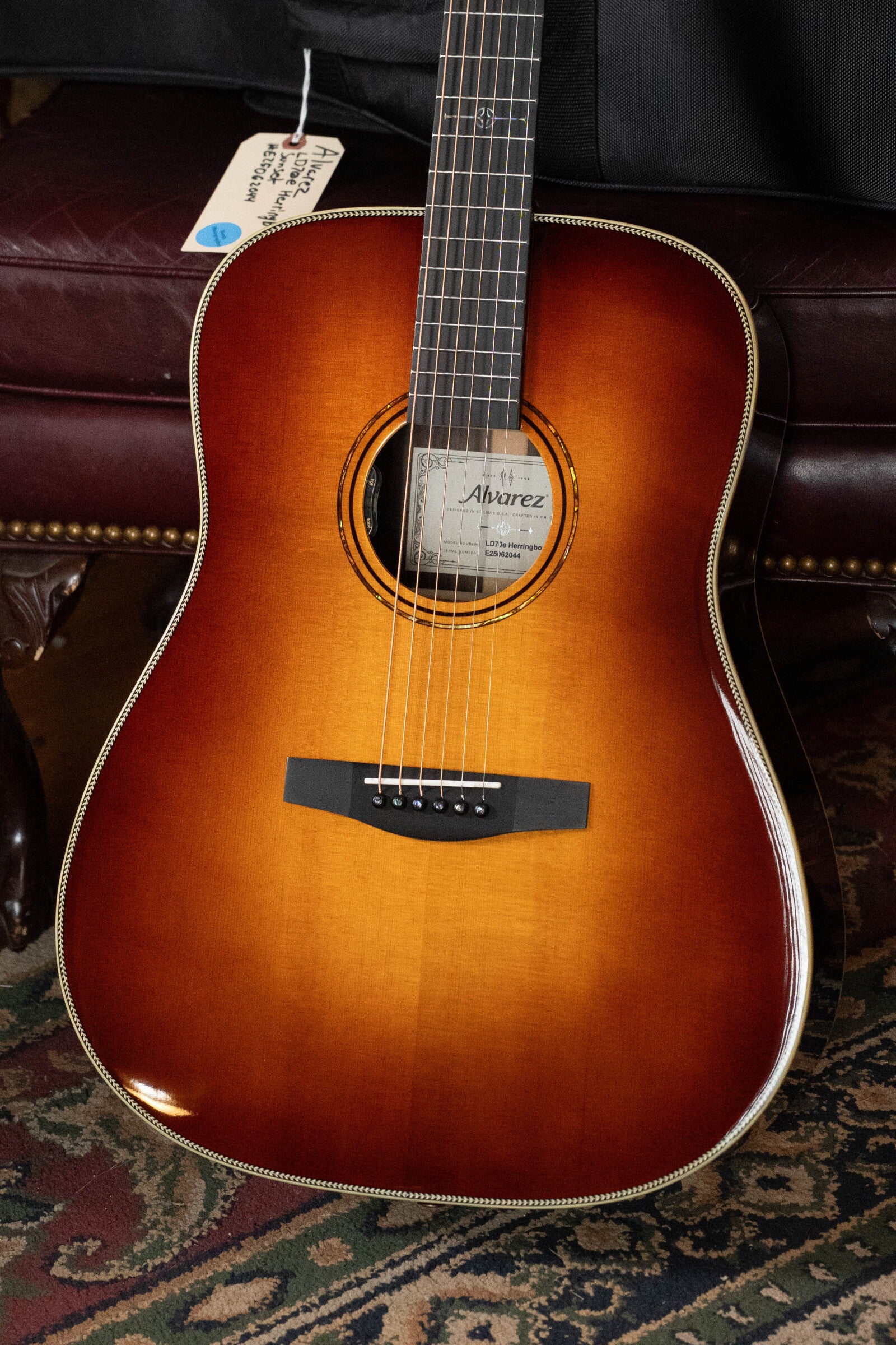 An Alvarez Laureate Series LD70e Herringbone Sunset Sitka/Rosewood Dreadnought Guitar w/LR Baggs Hifi Pickup #2044 rests on a patterned rug before a leather chair, with the Alvarez label visible inside the soundhole.