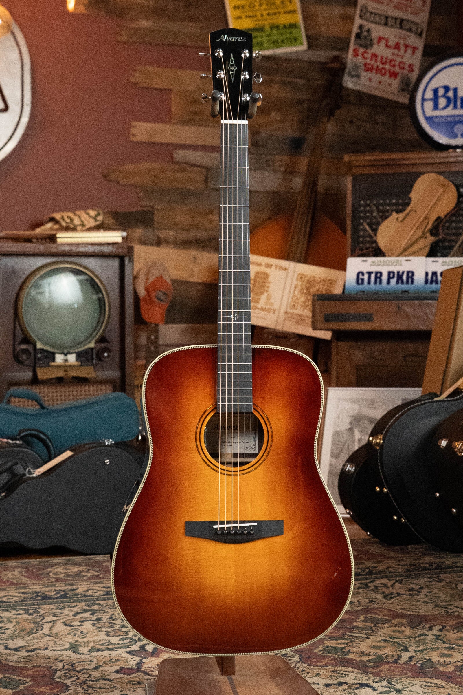 An Alvarez Laureate Series LD70e Herringbone Sunset Sitka/Rosewood Dreadnought Guitar w/LR Baggs Hifi Pickup stands upright in a cozy, vintage-themed room with wood-paneled walls, patterned rug, retro TV, guitar cases, and music posters.