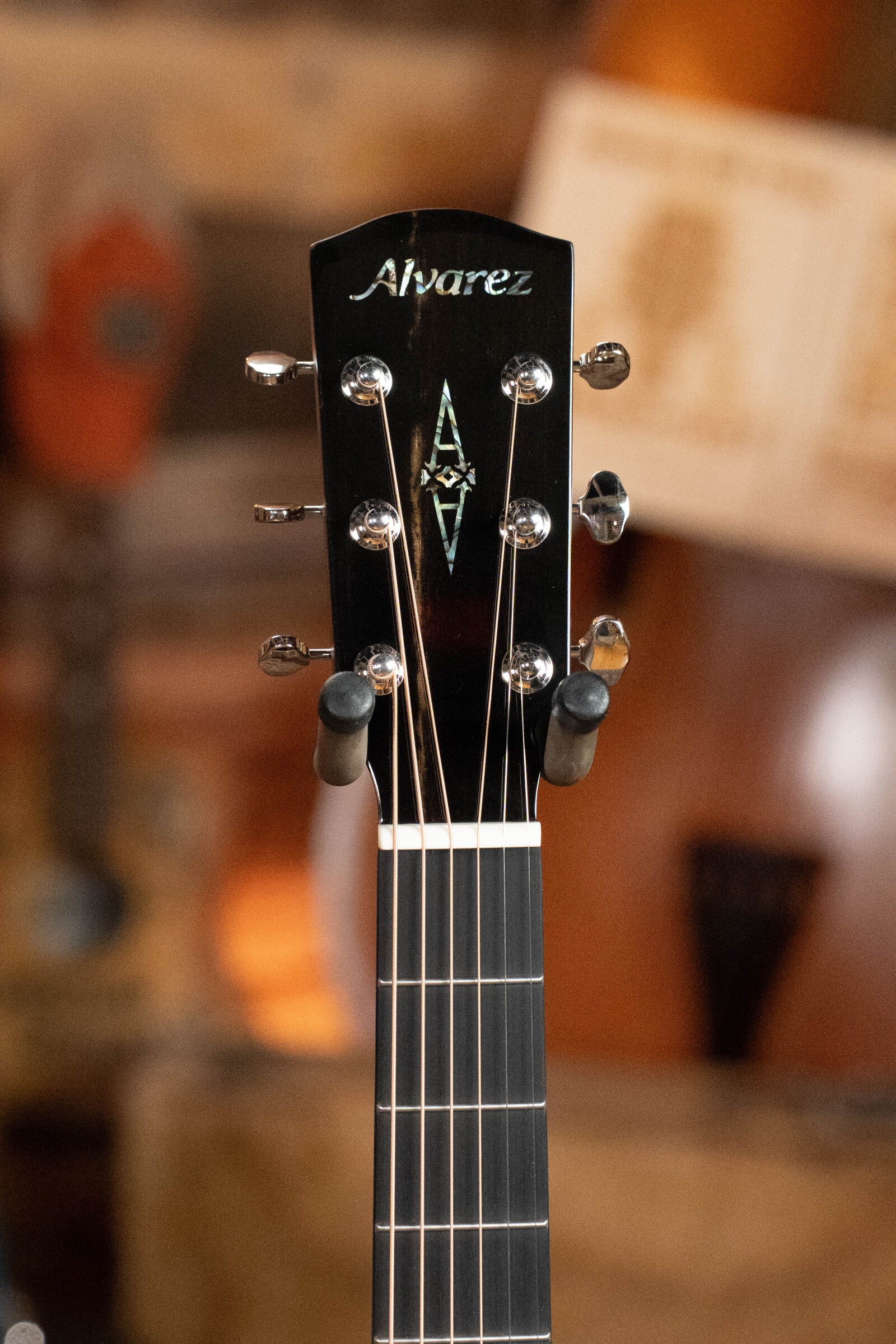 Close-up of the Alvarez Laureate Series LD70e Herringbone Sunset Sitka/Rosewood Dreadnought Guitar (#2044) headstock, highlighting tuning pegs, inlay, and Alvarez logo, with a blurred backdrop of other stringed instruments.