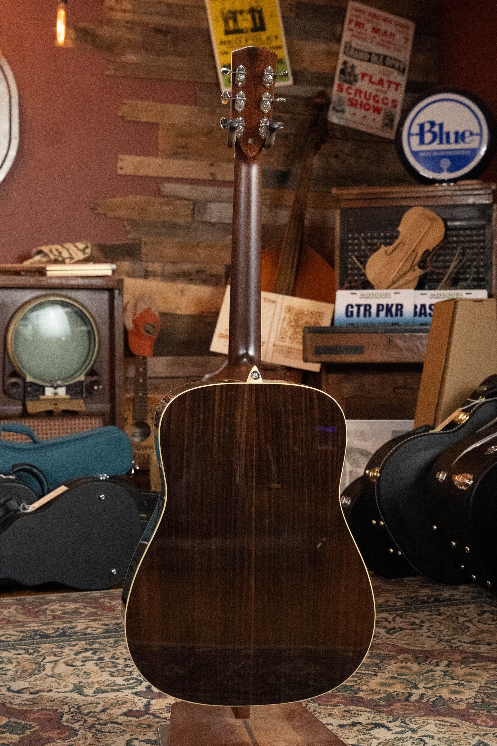 An Alvarez Laureate Series LD70e Herringbone Sunset Sitka/Rosewood Dreadnought Guitar w/LR Baggs Hifi Pickup (#2044) stands upright with its back facing forward in a cozy, vintage-style room filled with musical gear.