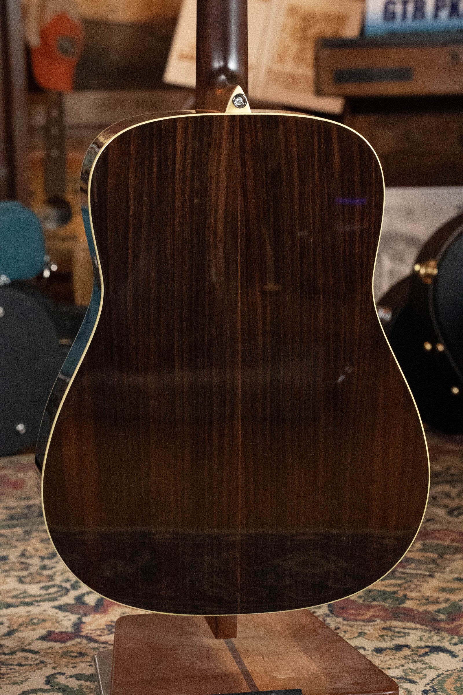 A close-up of the back of an Alvarez Laureate Series LD70e Herringbone Sunset Sitka/Rosewood Dreadnought Guitar w/LR Baggs Hifi Pickup, displayed upright on a stand in a music room with a patterned rug and instruments in the background.