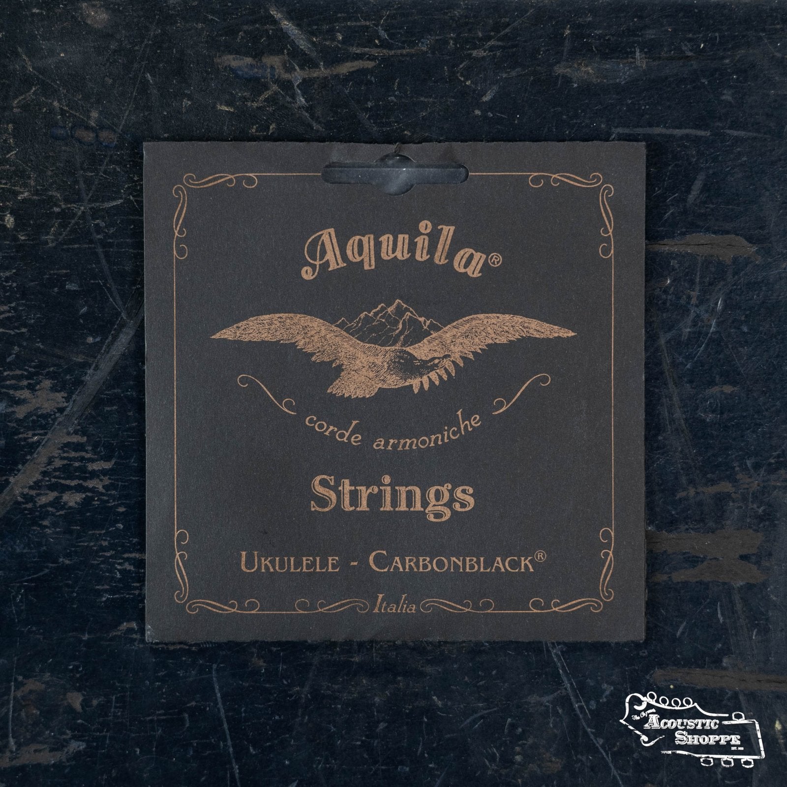 Aquila Carbonblack (141U) Soprano Ukulele Strings by Aquila, featuring gold lettering and an eagle logo on a dark background, are displayed on a worn black surface.
