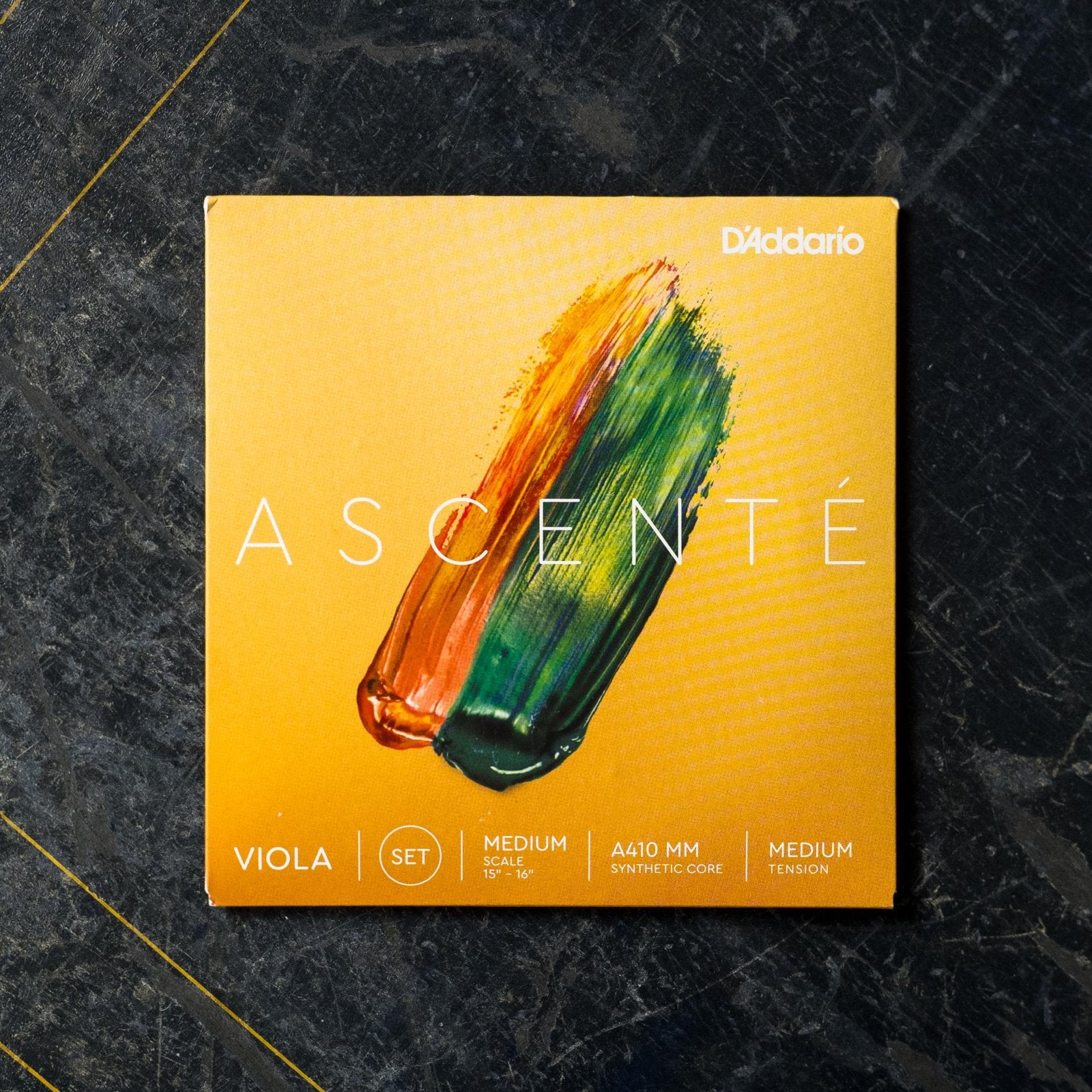 A DAddario Ascente A410 MM Medium Scale Synthetic Core Viola Strings set by DAddario, in packaging with orange and green paint strokes on yellow, labeled Viola Set, Medium Scale, Medium Tension, on a dark textured background.