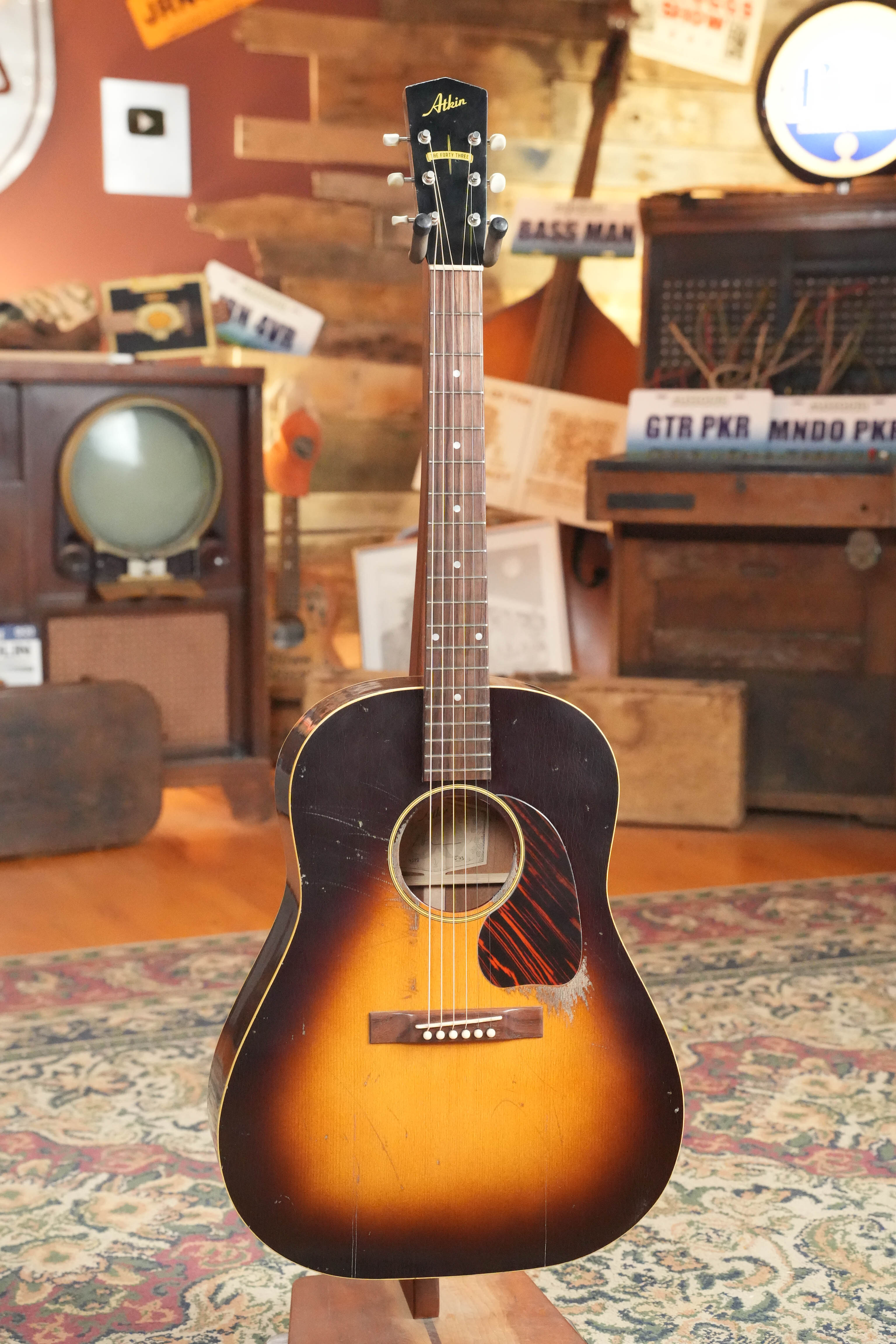 An Atkin Guitars J43 45MM Baked Sitka/Mahogany Heavy Aged Slope Shoulder Dreadnought Acoustic Guitar (#4398) stands upright on a stand in a cozy retro room filled with vintage decor, patterned rug, old TV, radio, and signs.