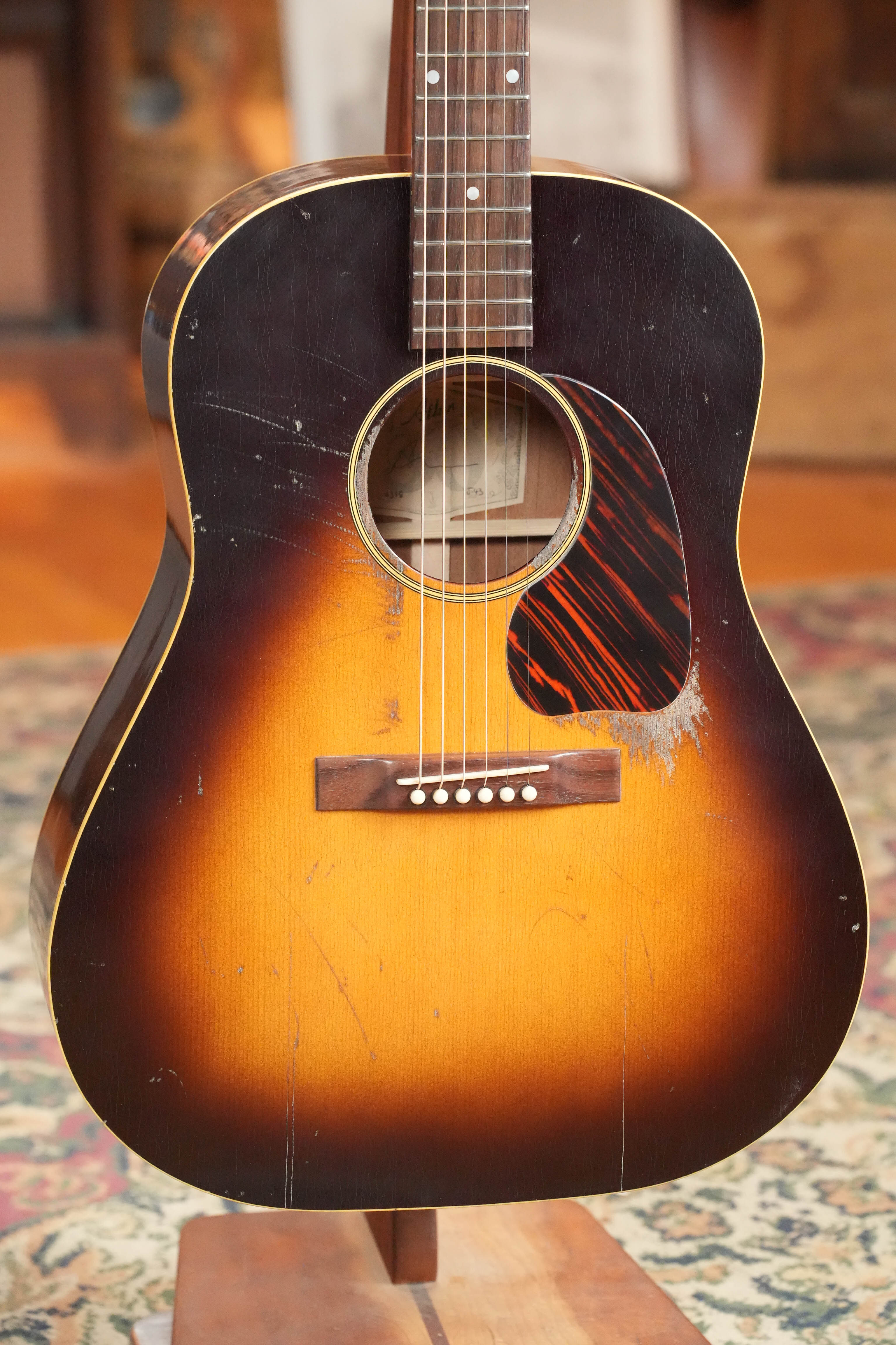 The Atkin Guitars J43 45MM Baked Sitka/Mahogany Heavy Aged Slope Shoulder Dreadnought Acoustic Guitar #4398, with a sunburst finish, tortoiseshell pickguard, and visible wear, stands upright on a patterned rug.