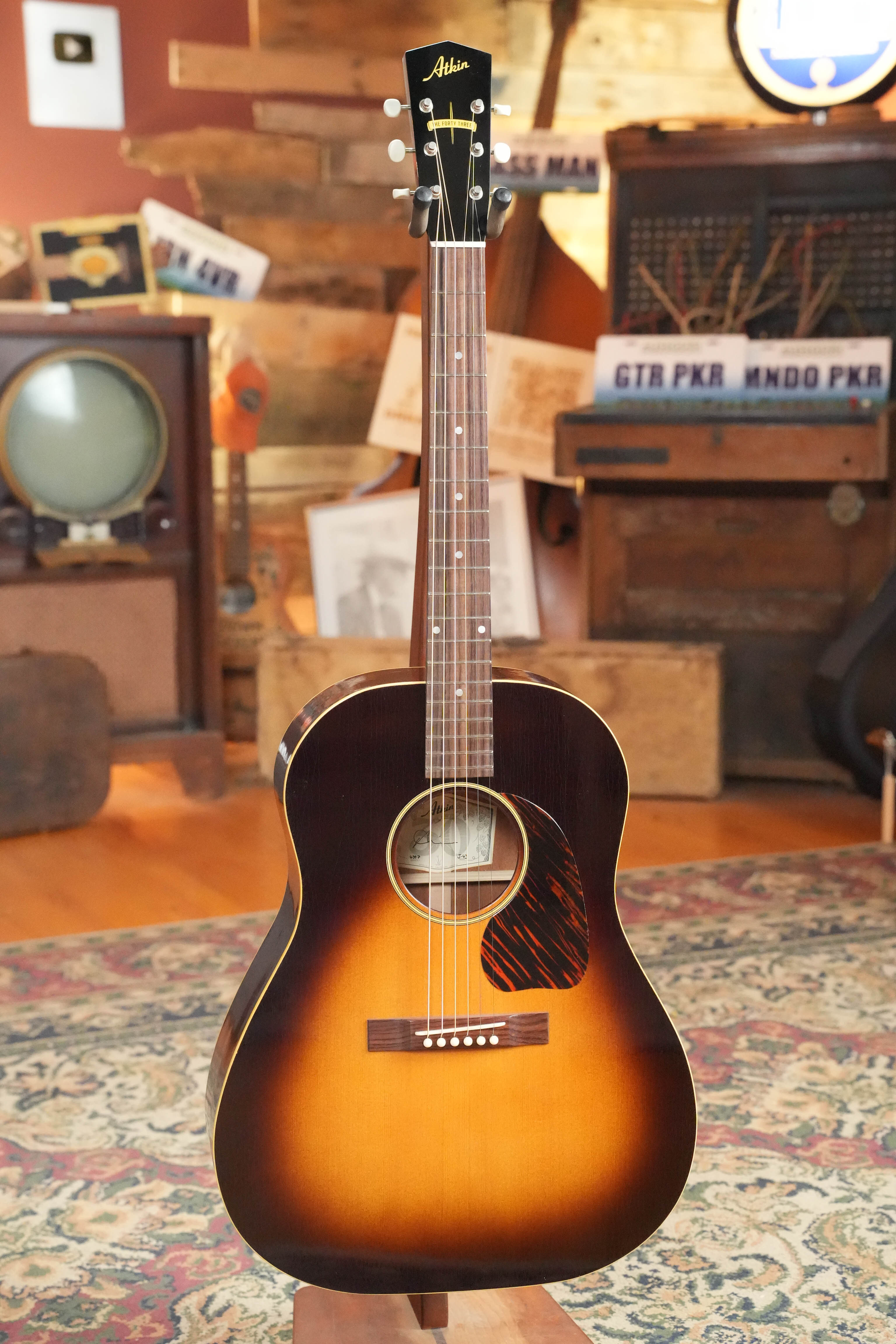An Atkin J43 Baked Sitka/Mahogany Heavy Aged Slope Shoulder Dreadnought Acoustic Guitar by Atkin Guitars, with a vintage sunburst and aged nitro finish, stands on a patterned rug amid retro electronics, wood accents, and vintage decor.