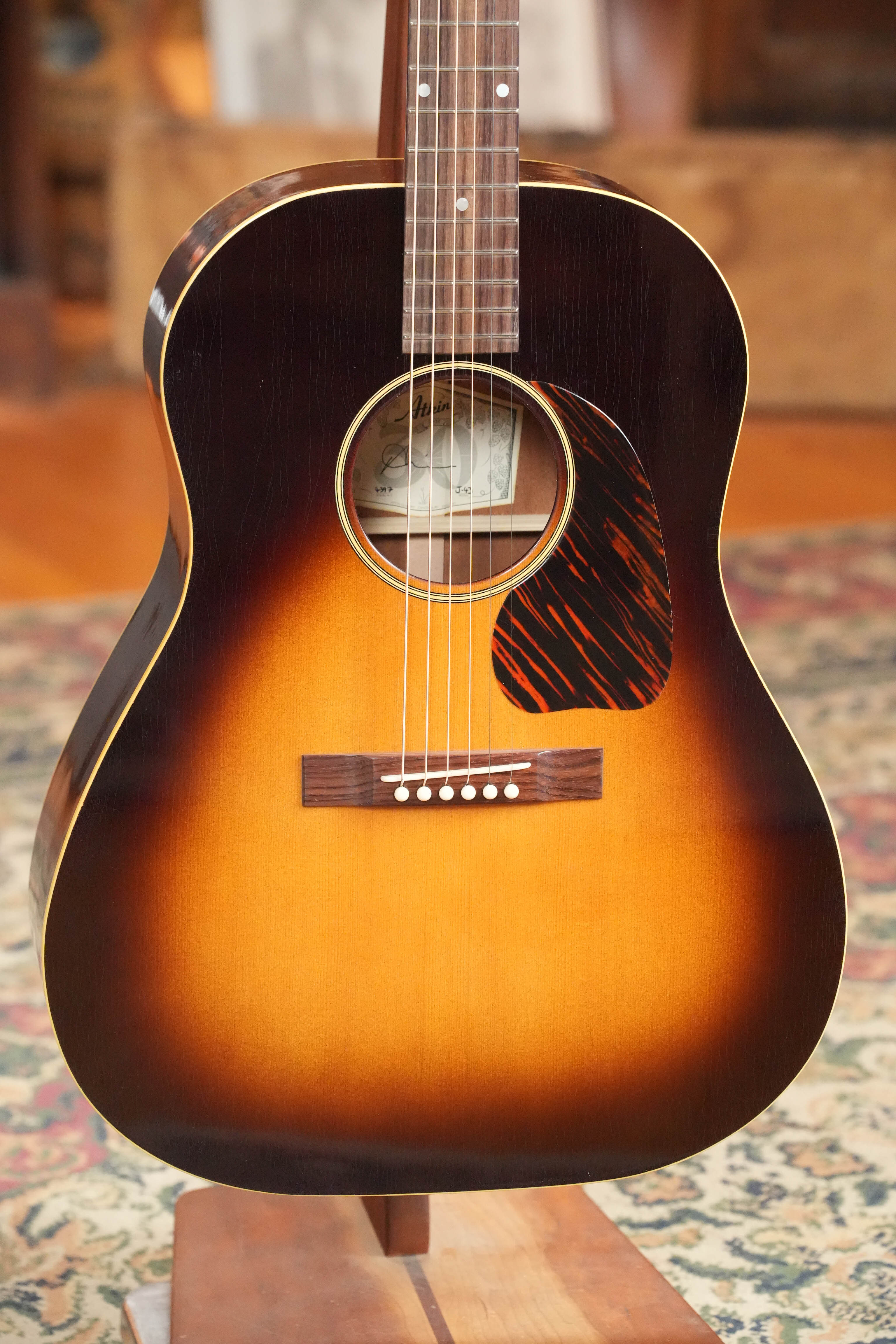 The Atkin J43 Baked Sitka/Mahogany Heavy Aged Slope Shoulder Dreadnought Acoustic Guitar #4397 by Atkin Guitars, featuring a sunburst finish and tortoiseshell pickguard, stands upright before a patterned rug and wooden furniture backdrop.