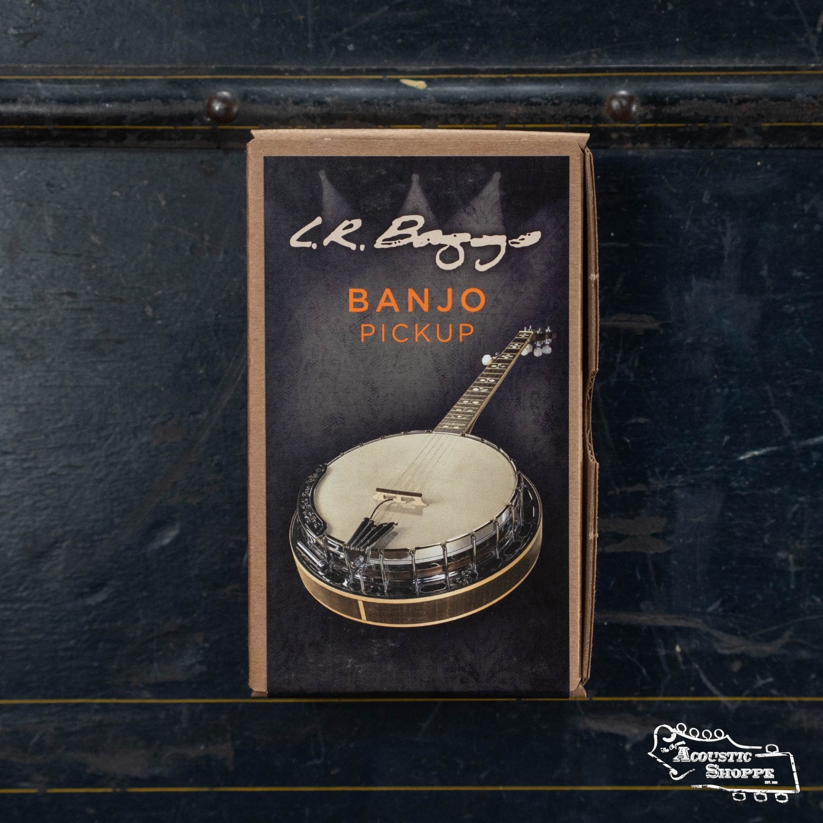 A box of the L.R. Baggs Banjo Pickup 5/8, featuring a banjo image and showcasing its bridge pickup design, sits on a worn wooden surface with The Acoustic Shoppe logo at the bottom right.