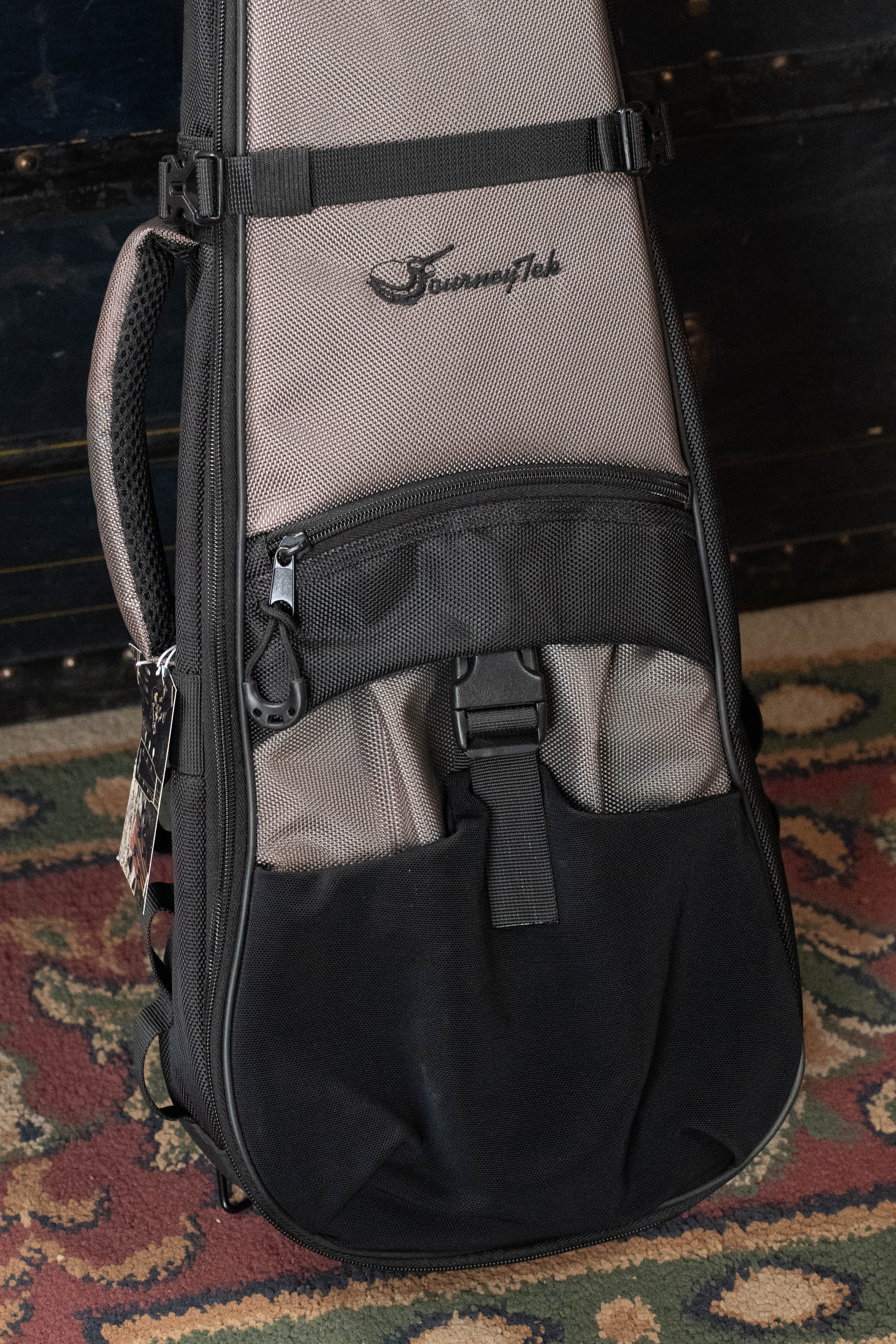 A gray and black Journey Instruments JourneyTek Concert Ukulele Case BG002 with padded protection sits upright on a patterned rug, featuring a zippered pocket and handle for easy carrying.