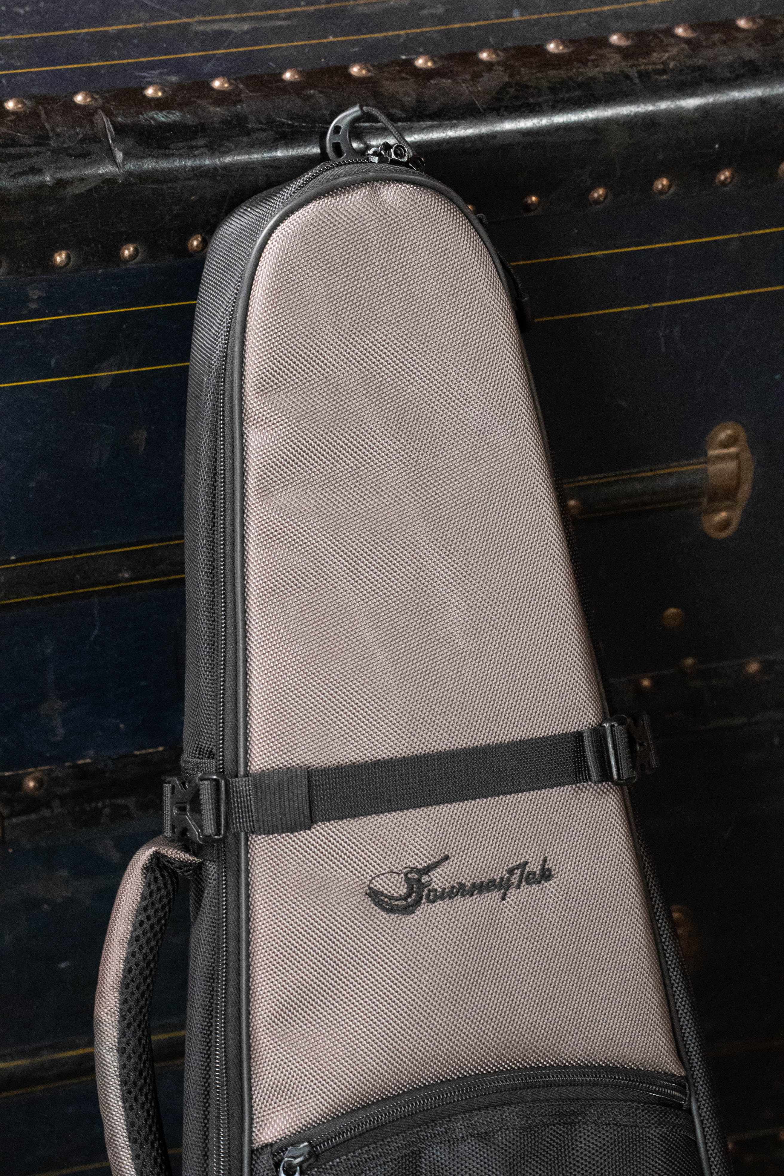 The Journey Instruments JourneyTek Concert Ukulele Case BG002 in gray and black hangs in front of a vintage trunk, showcasing stylish protection for your instrument with the distinct TourenTek logo.