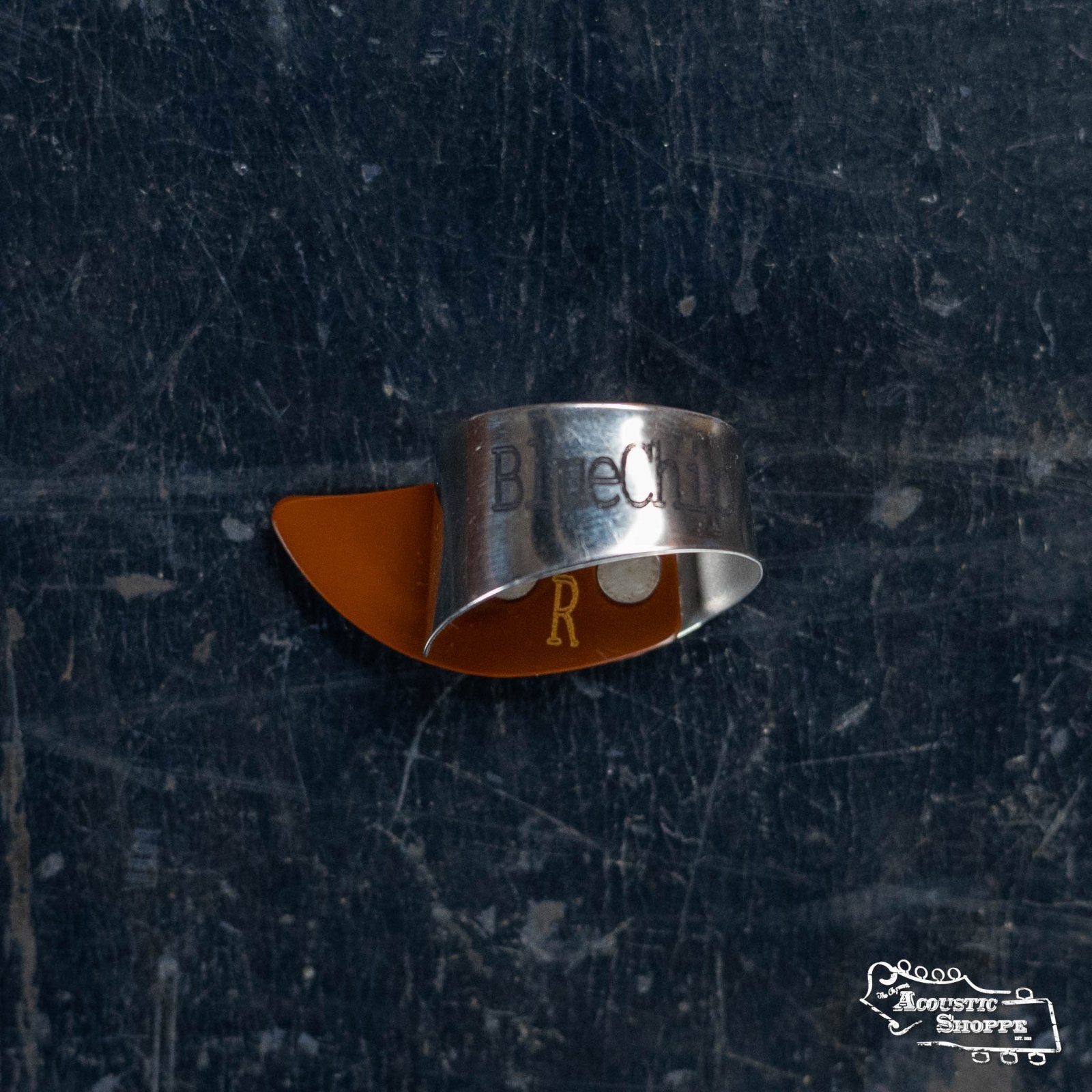 A BlueChip Picks (BCT-BRL) thumbpick and a brown banjo thumb pick rest on a worn, dark surface, with The Acoustic Shoppe logo in the bottom right corner.