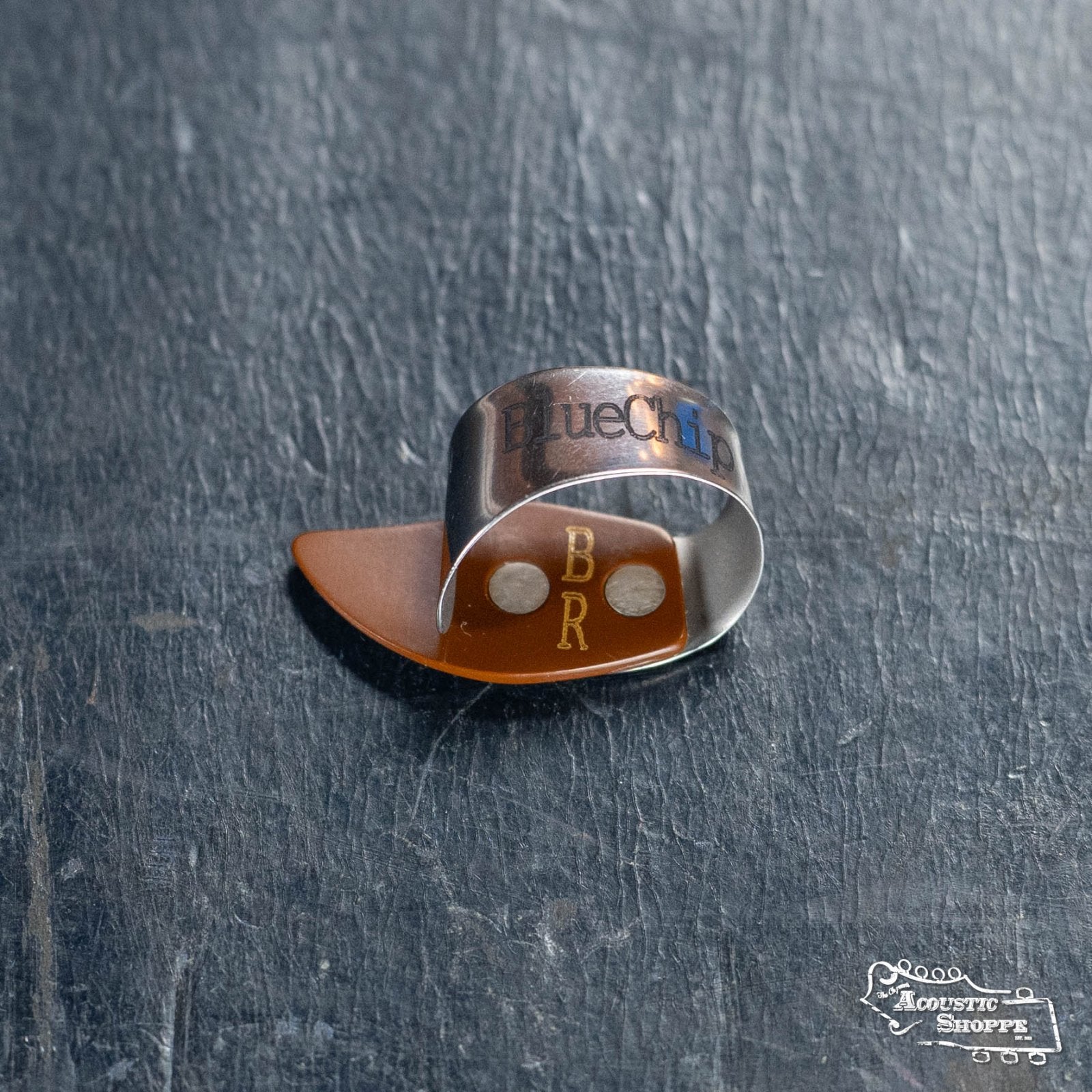 A BlueChip Picks (BCT-BRL) thumbpick by BlueChip, made of silver metal, sits on a dark surface. An orange plastic thumb pick labeled BR is inside it, and The Acoustic Shoppe logo appears in the lower right corner.