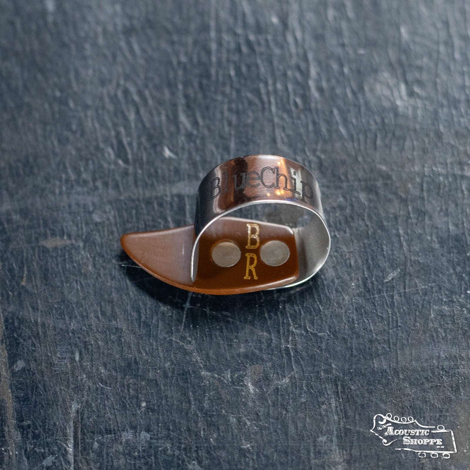 The BlueChip Picks (BCT-BRM) thumbpick by BlueChip, featuring B R engraved on plastic and Blues on the metal, sits on a dark textured surface. The Acoustic Shoppe logo appears in the lower right—a stylish banjo accessory.