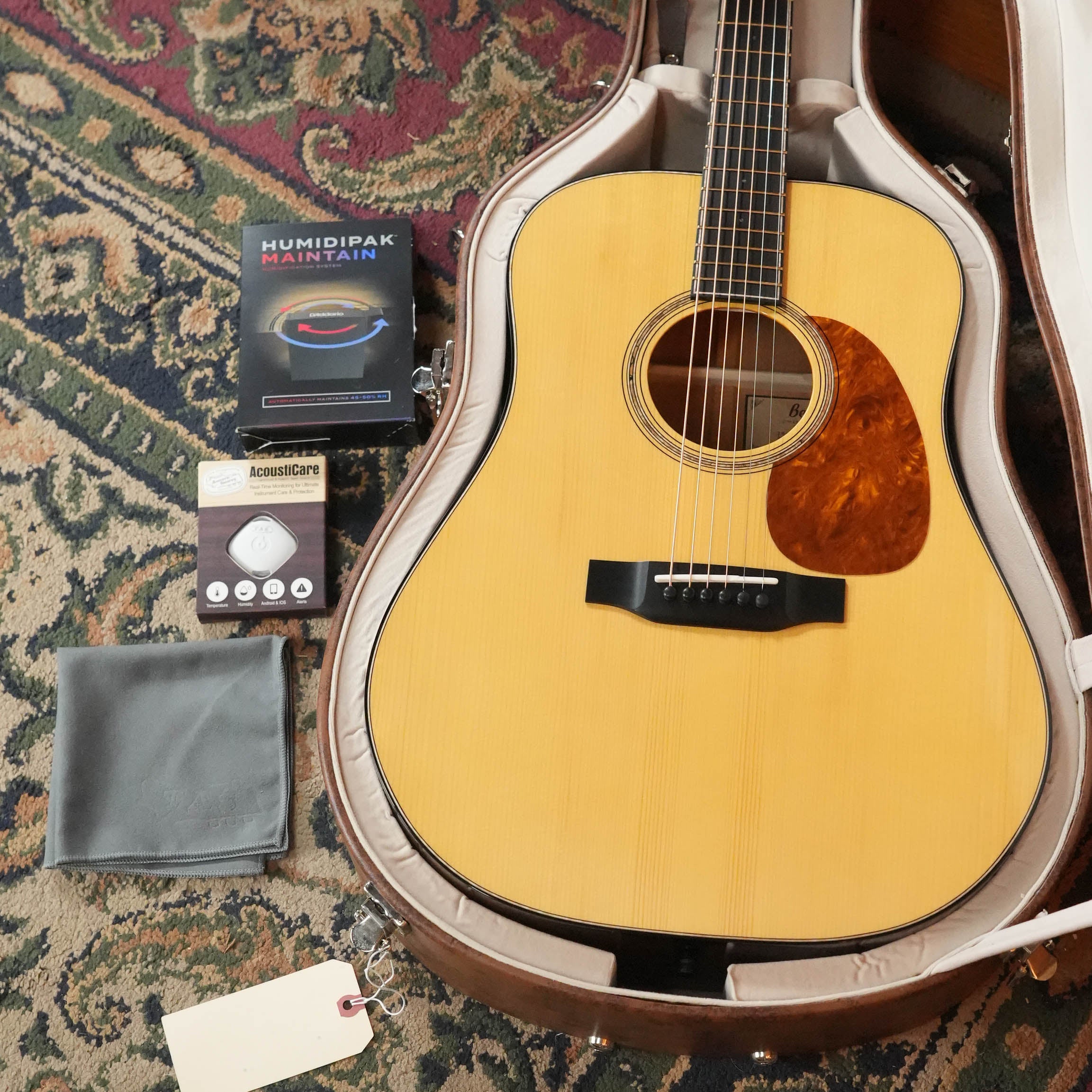 A Bedell Guitars Special Edition 1940 Dreadnought Narrow Nut Acoustic Guitar #5004 rests in an open case on a patterned rug, with a gray cloth, D’Addario Humidipak Maintain kit, AccessCare box, and blank white tag nearby.