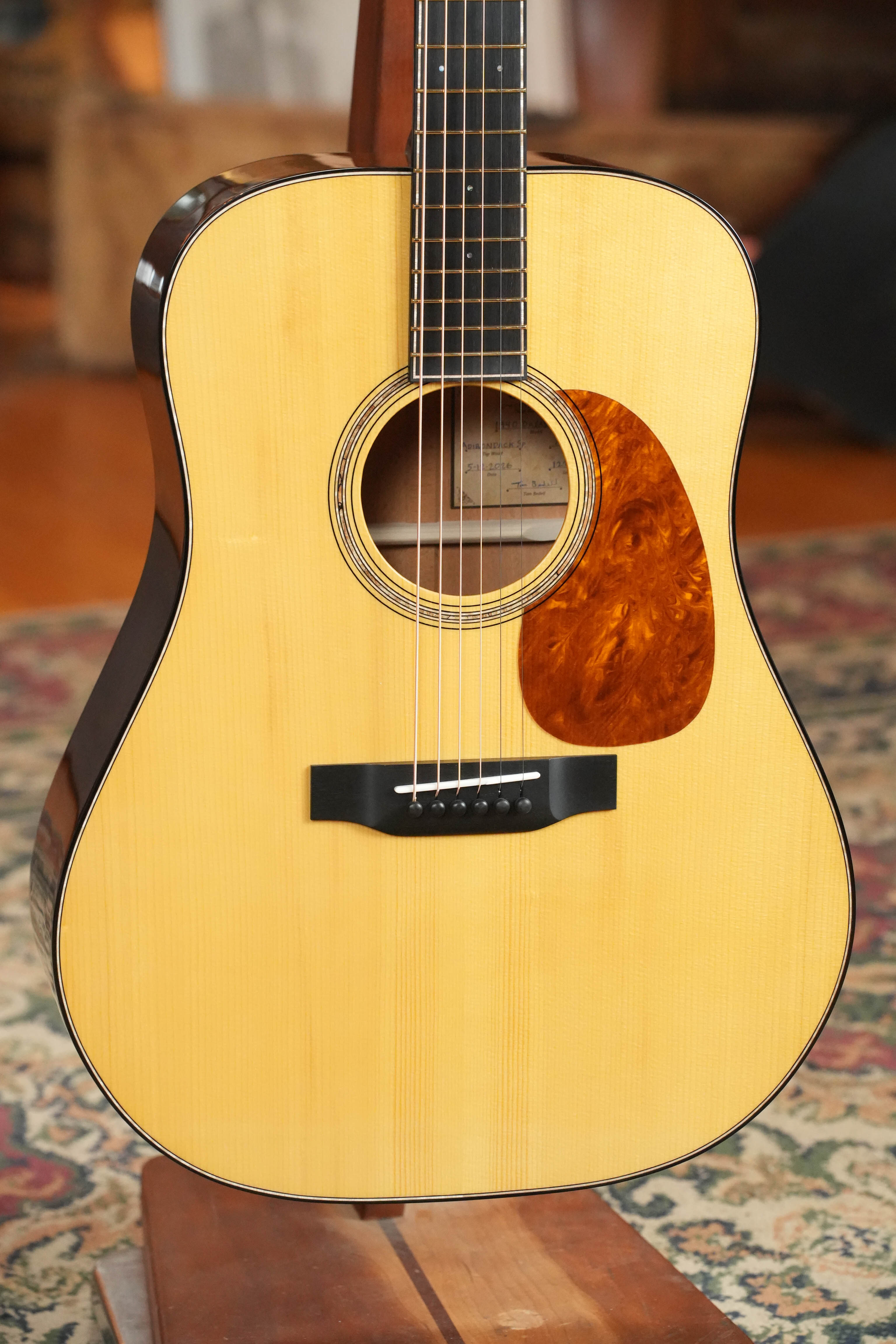 A close-up of the Bedell Guitars Special Edition 1940 Dreadnought Narrow Nut Acoustic Guitar #5004, featuring an Adirondack spruce top and tortoiseshell pickguard, resting on a stand in a warmly lit room with a patterned rug.