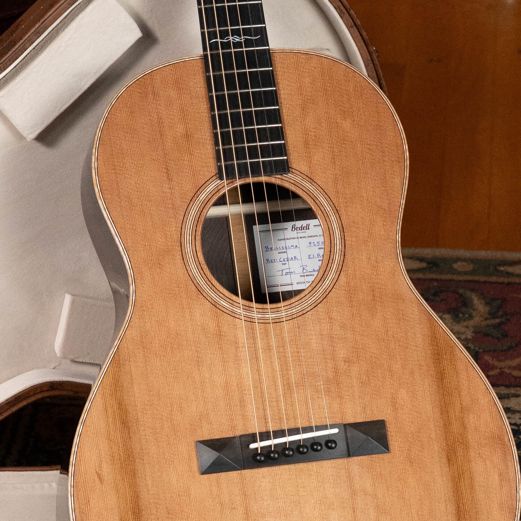 A Bedell Angelica Bellissima Red Cedar/Rosewood Parlor Guitar w/K&K Pure Mini Pickup (#5002) by Bedell Guitars, with a natural wood finish, rests in an open white case; its label is visible through the soundhole.