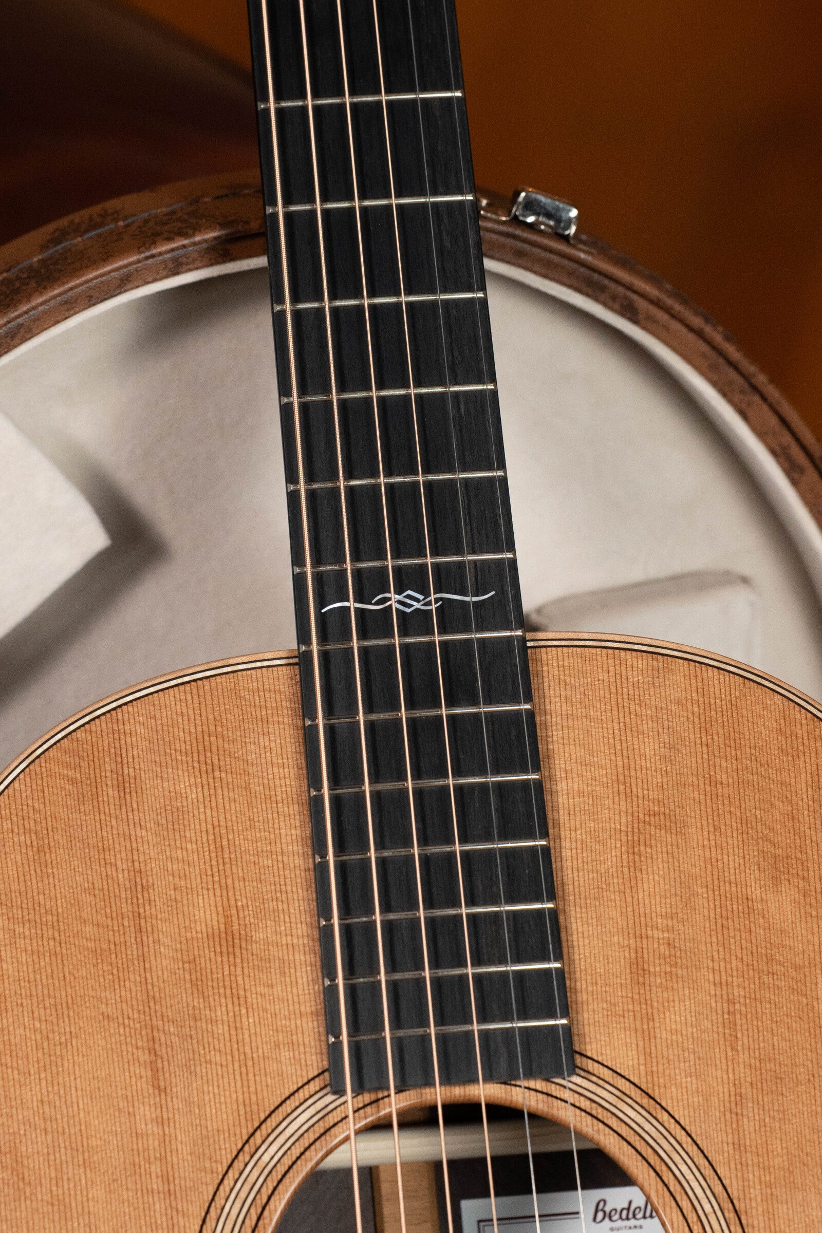 Close-up of the Bedell Angelica Bellissima Red Cedar/Rosewood Parlor Guitar’s fretboard and sound hole, showing detailed wood grain and inlay design, with part of a banjo in the background. Features a K&K Pure Mini Pickup. Brand: Bedell Guitars.